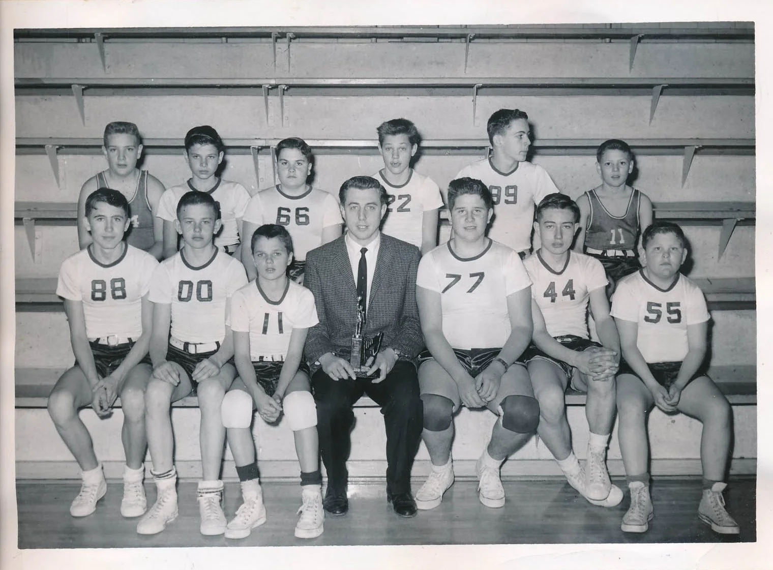 A vintage black-and-white photo of a boys' basketball team with their coach, seated and standing on gym bleachers. The boys are wearing numbered jerseys and the coach is in a suit, holding a trophy.