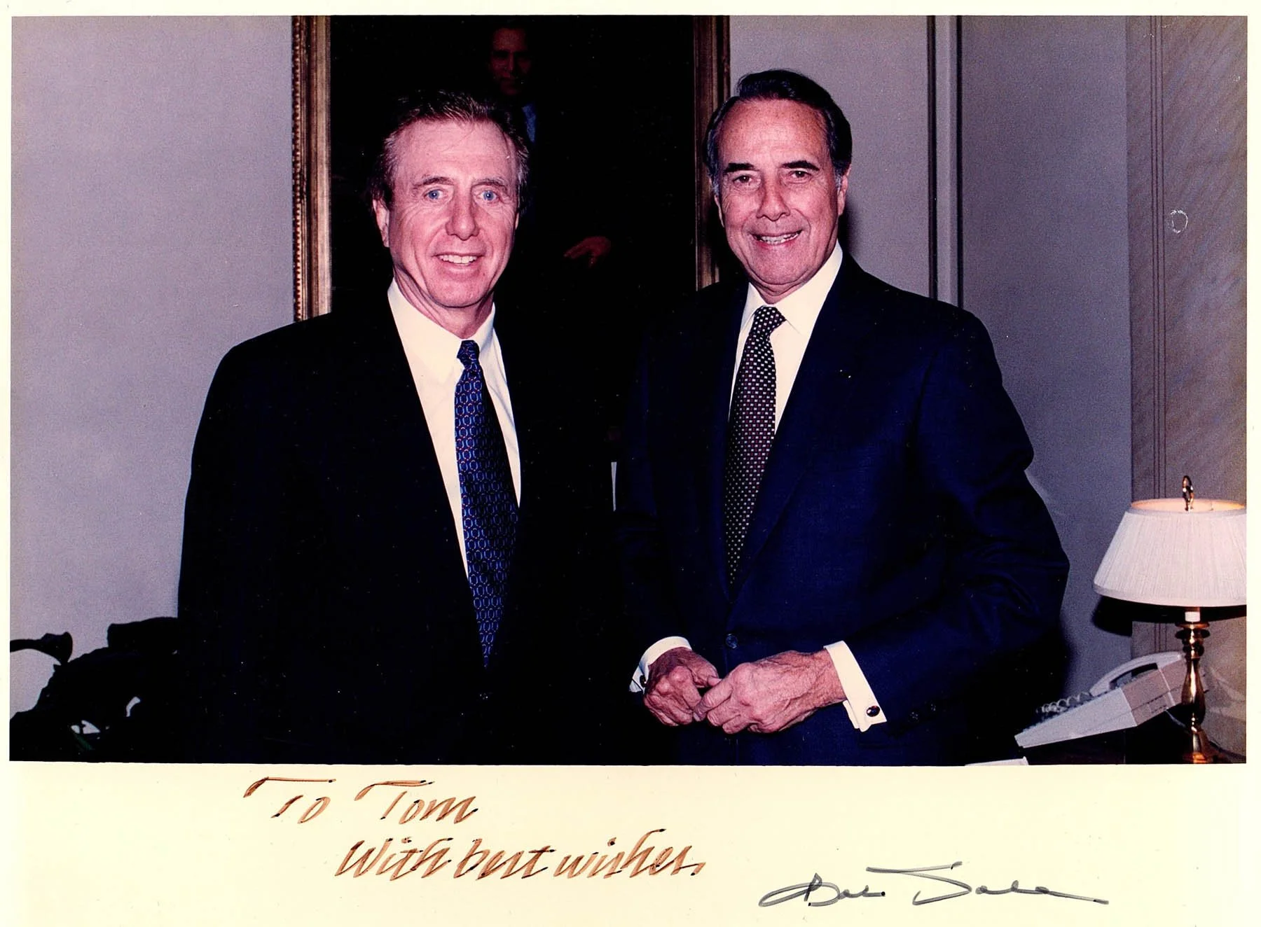 Two men in business suits standing in an office, smiling, with a framed portrait behind them and a desk with a lamp and phone to the right.