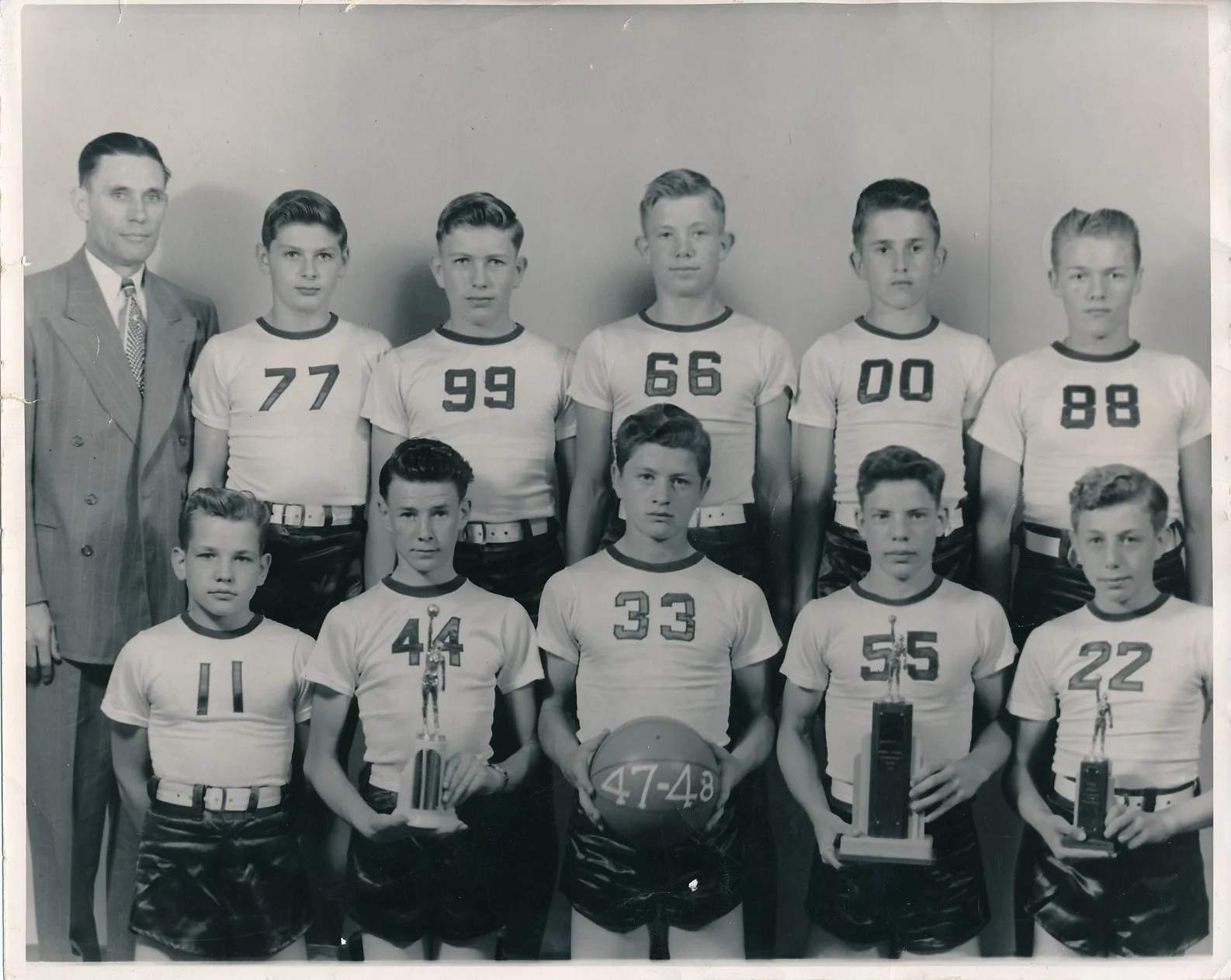 A black and white photo of a boys' basketball team with their coach. The team is arranged in two rows, with five boys standing in the back row and four boys kneeling in front. Each boy is wearing a uniform with a number on the front and holding troph