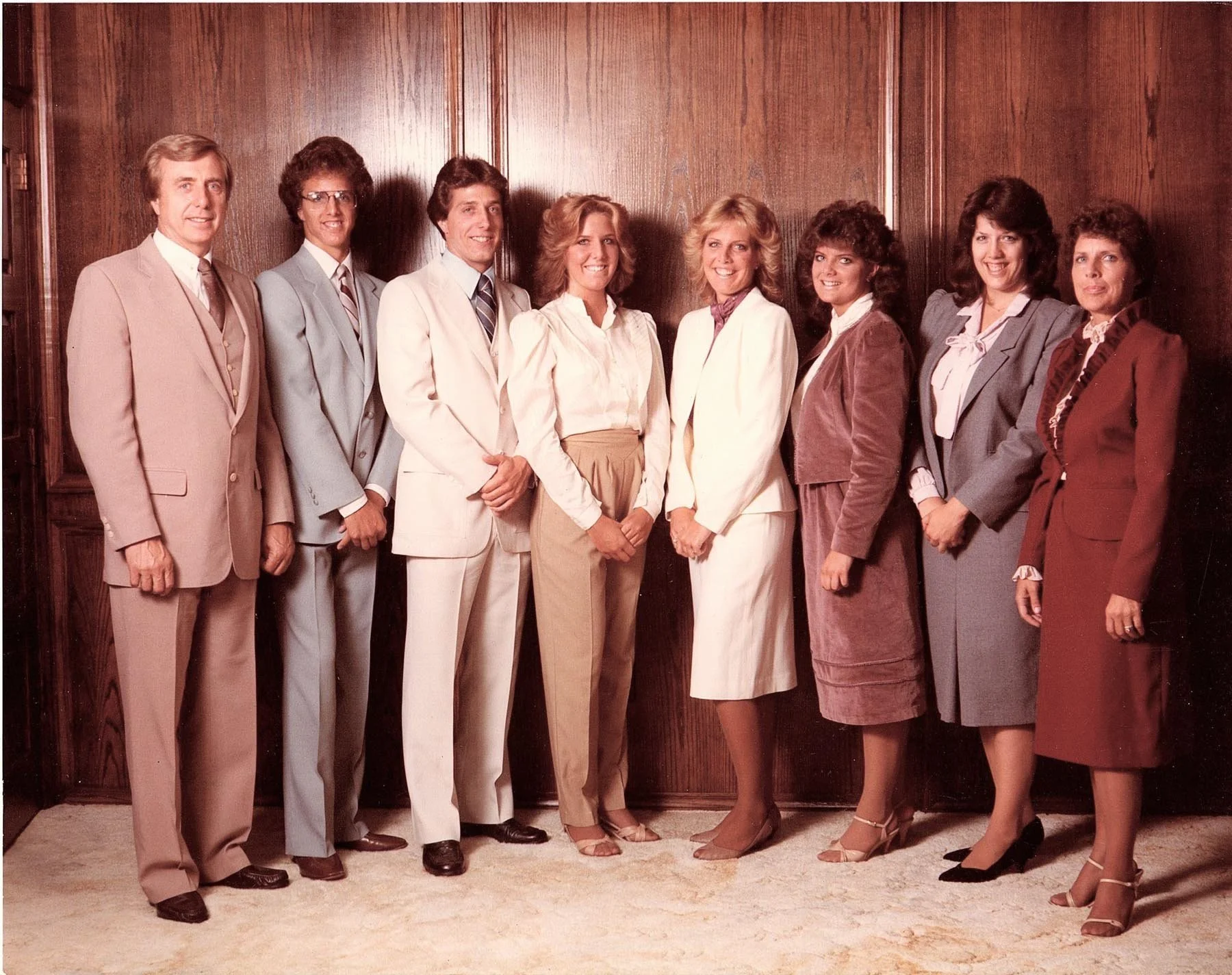 Group of nine professionally dressed individuals standing in a line indoors against a wooden wall, smiling at the camera.
