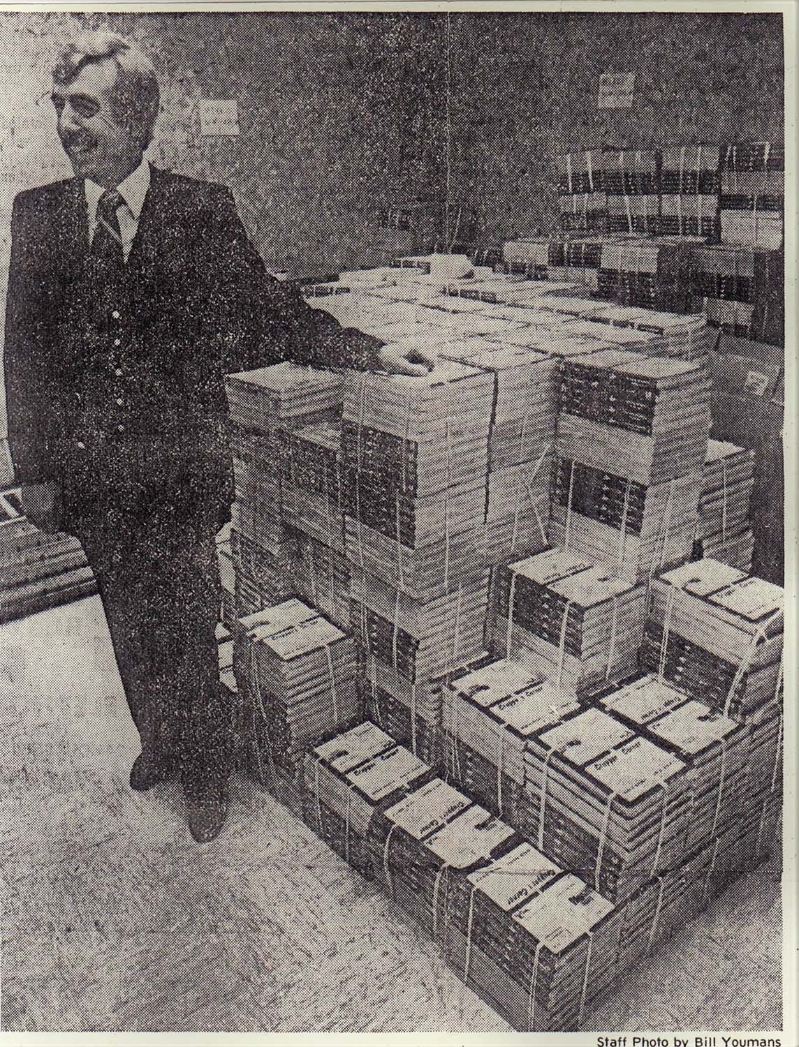 A man in a suit standing next to stacks of magazines or newspapers in an office setting.