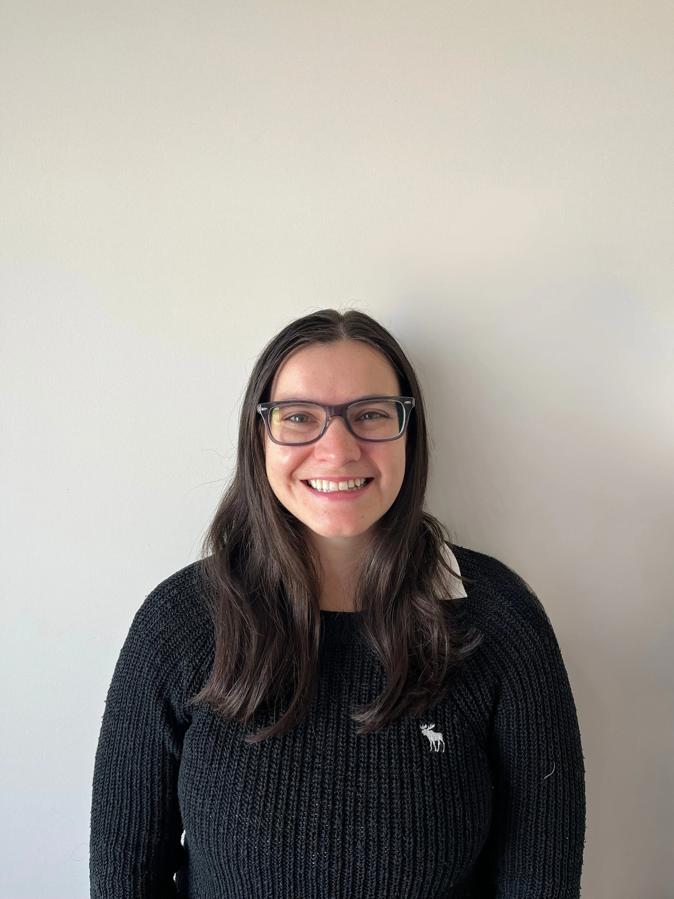 A young woman with long dark hair, glasses, and a black sweater smiling in front of a plain white wall.