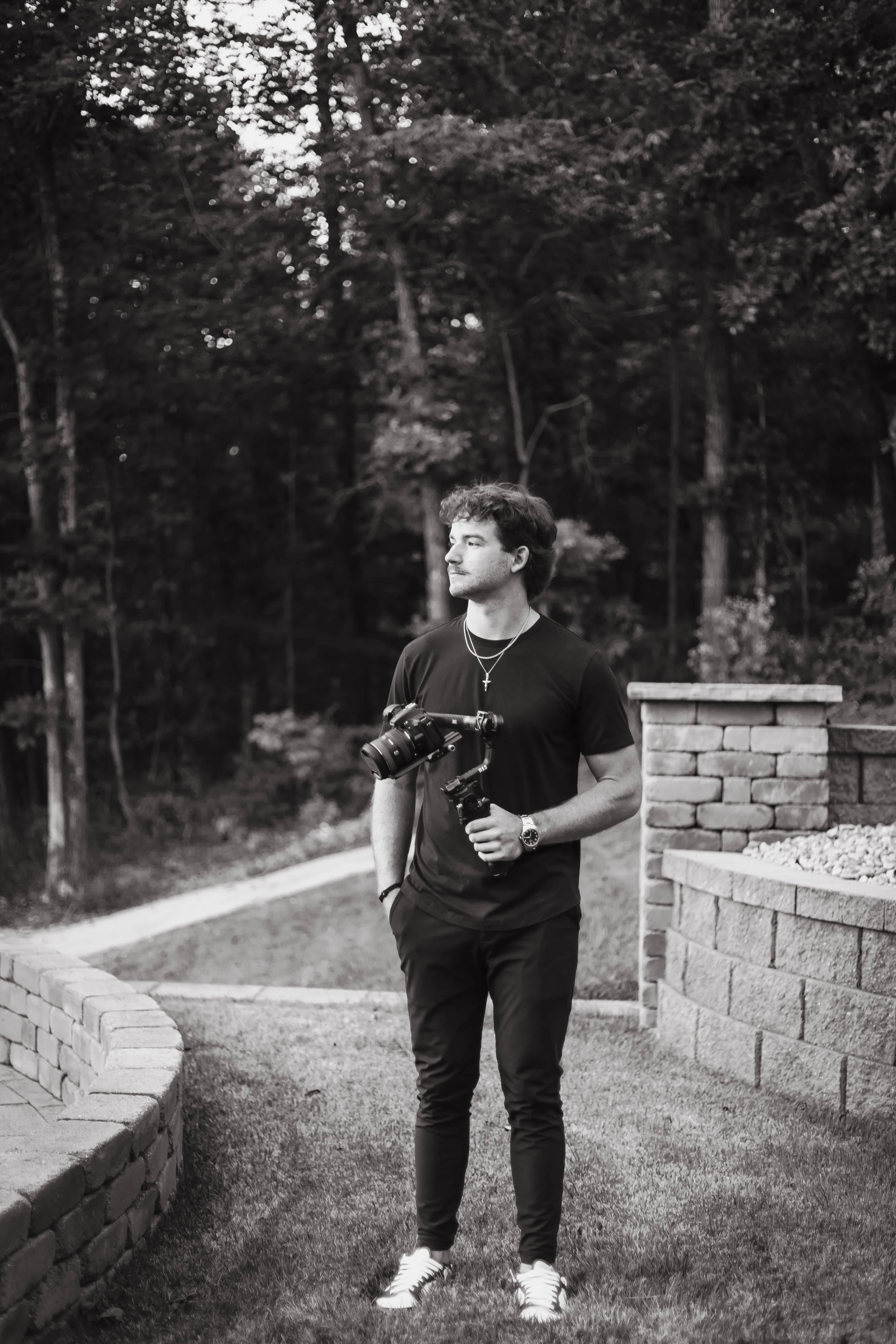 A young man with dark, curly hair, wearing a black t-shirt, black pants, sneakers, a watch, and necklaces, stands outdoors on a grassy area with a brick wall and forest in the background. He is holding a camera stabilizer with a camera attached.