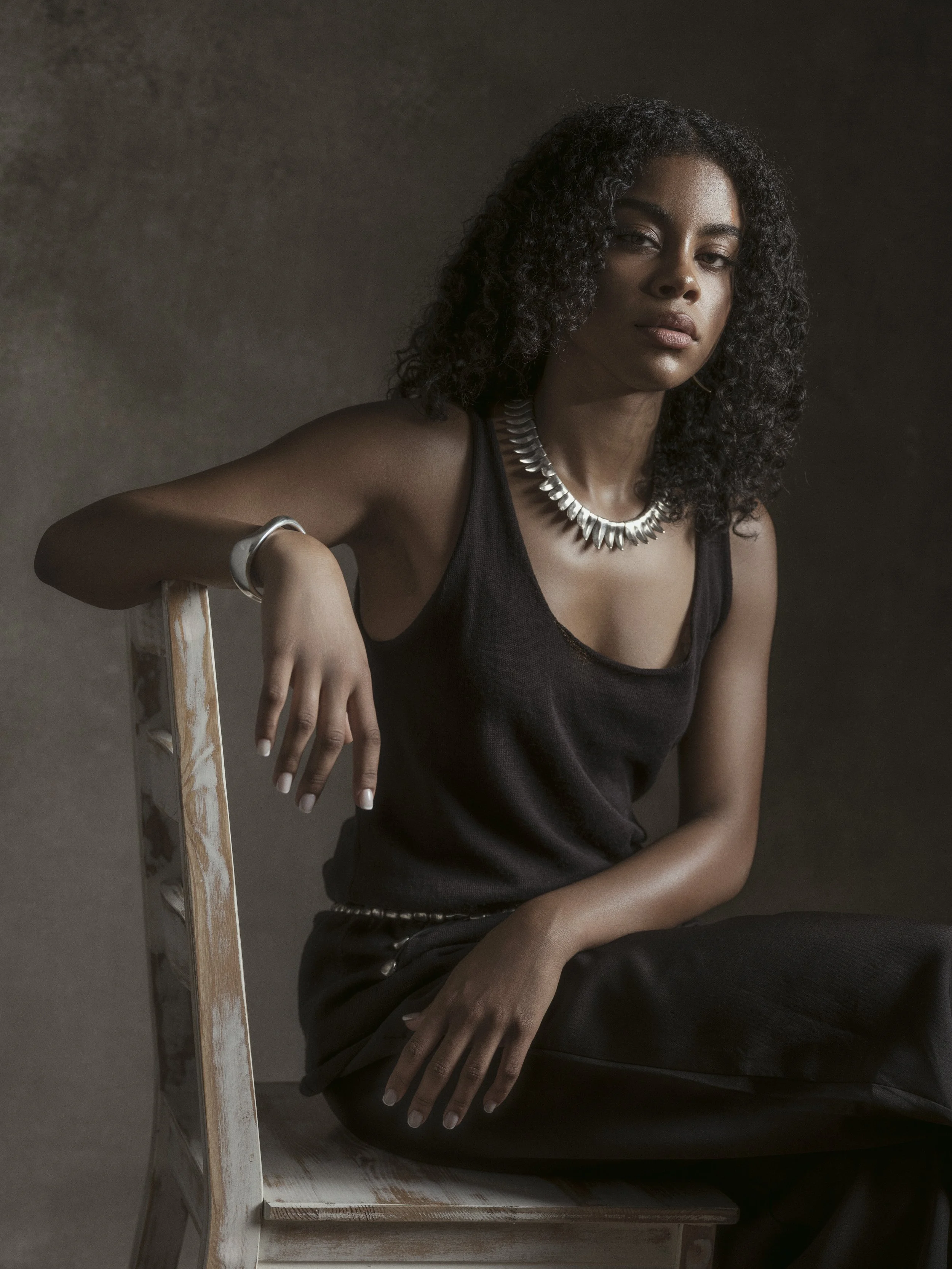A young woman with curly black hair seated on a white wooden chair, wearing a black sleeveless top, black pants, silver jewelry including a necklace, bracelet, and belt, poses with a neutral expression against a plain, textured background.