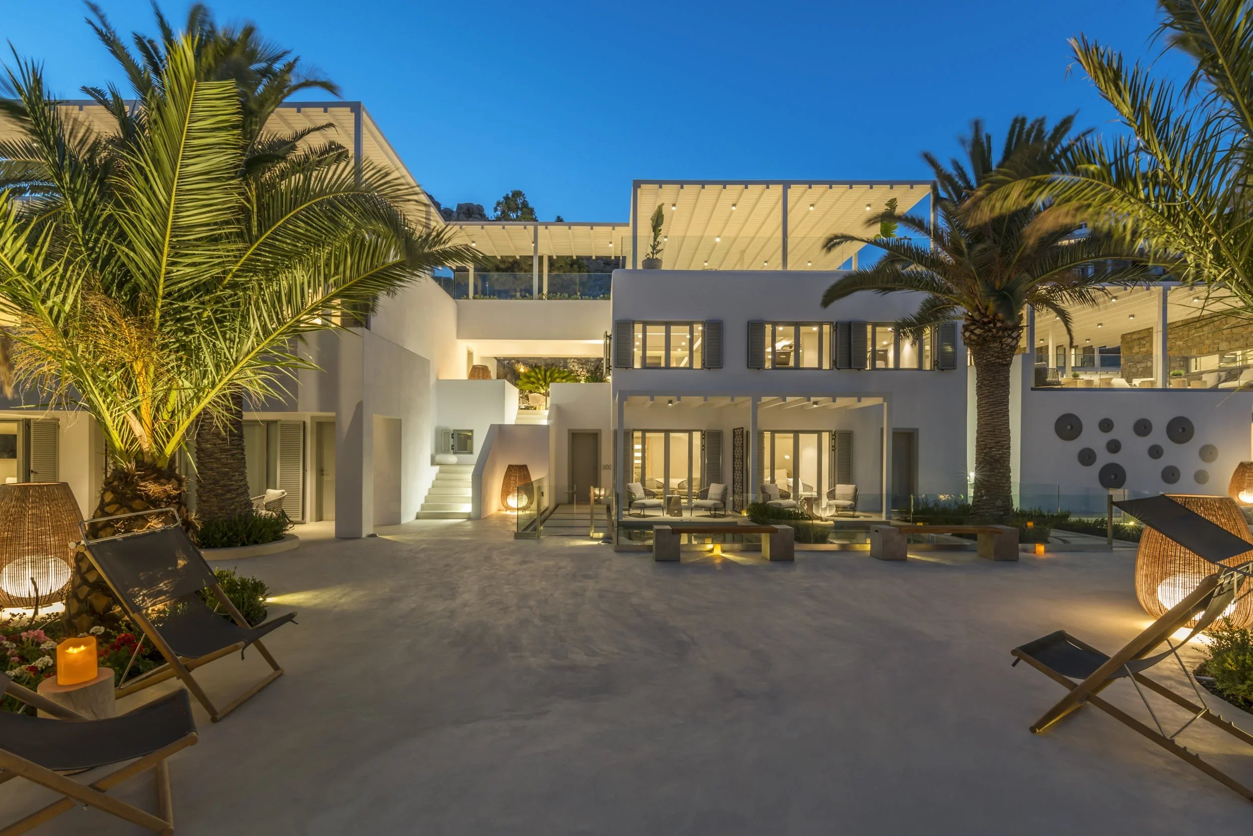 Luxury modern house with outdoor lounge area and palm trees at dusk, illuminated with ambient lighting.