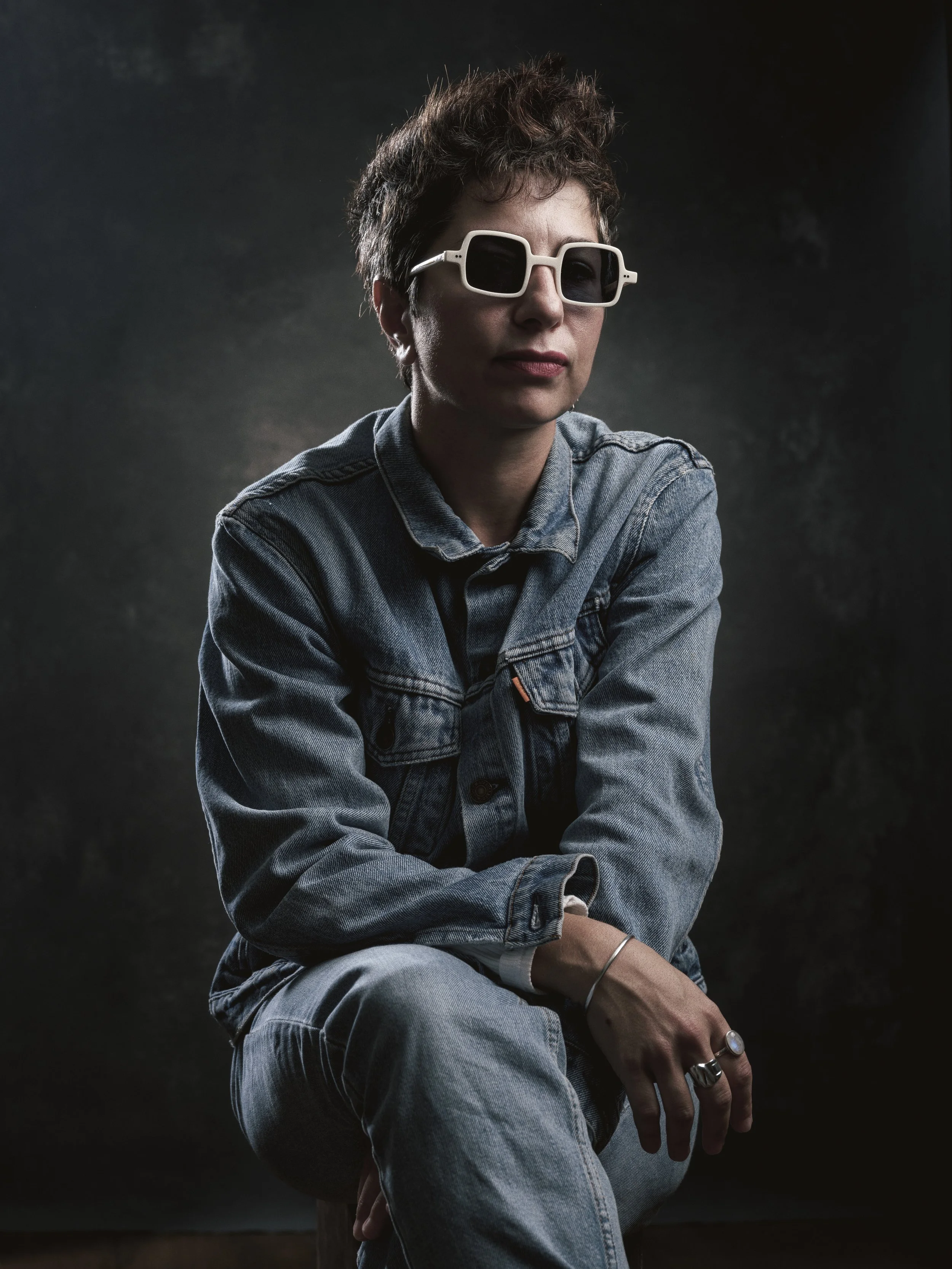 A person with short, curly hair wearing large, square sunglasses and a denim jacket, sitting with one knee up against a dark, textured background.