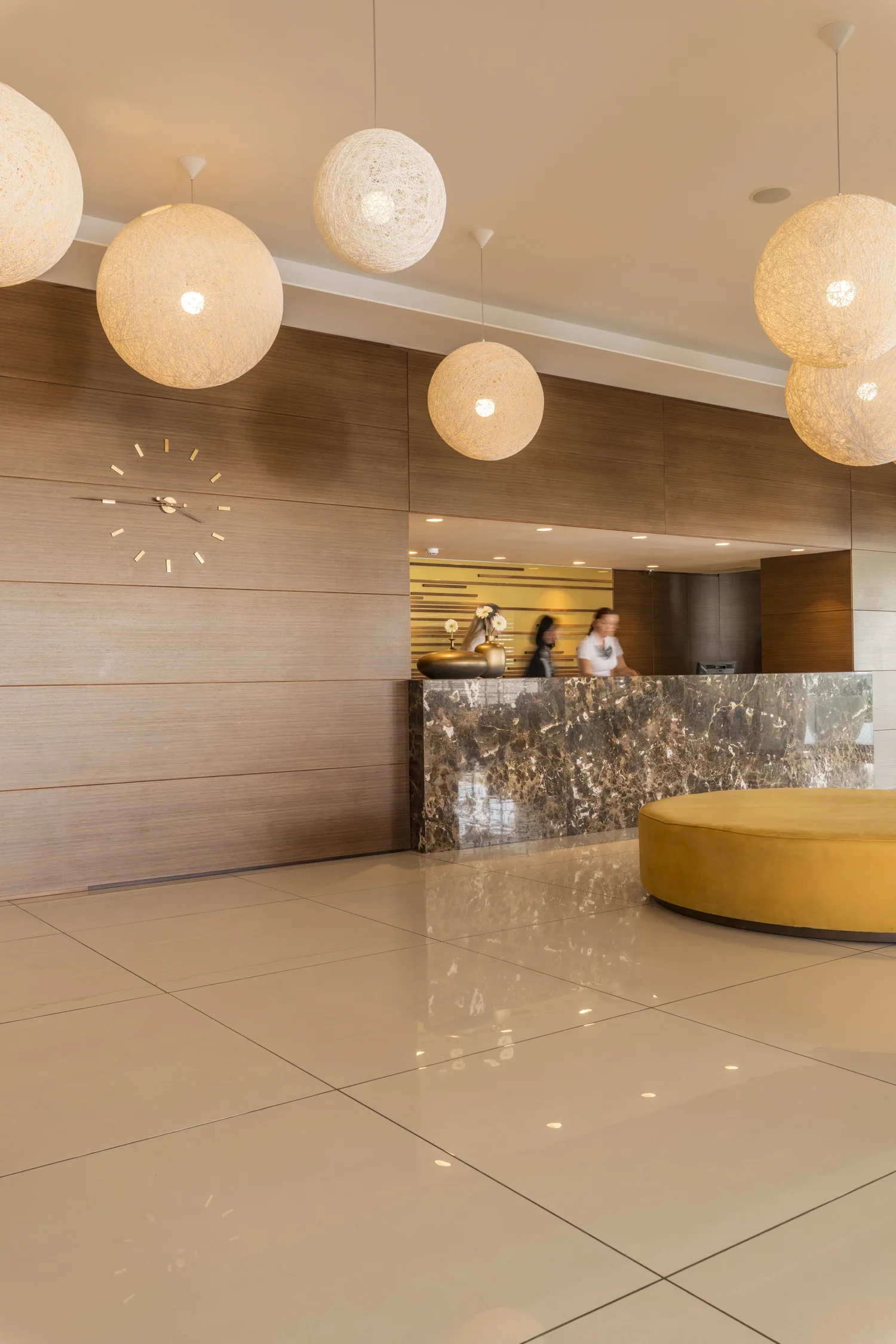 Modern hotel lobby with a reception desk, hanging spherical pendant lights, a yellow upholstered round bench, and women at the reception counter.