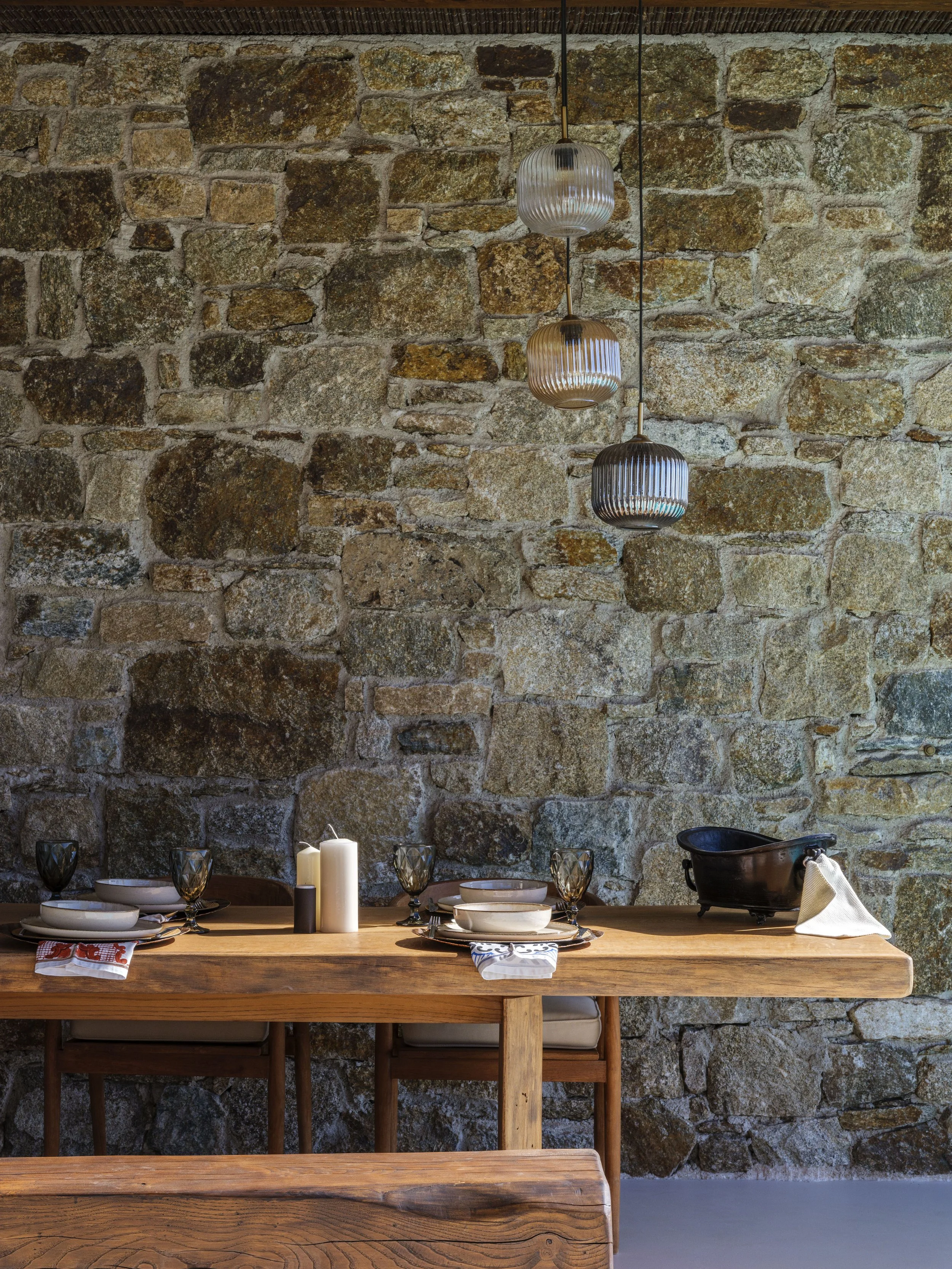 A rustic dining area with a stone wall background, a wooden table set for a meal with plates, glasses, and candles, and three hanging pendant lights above.