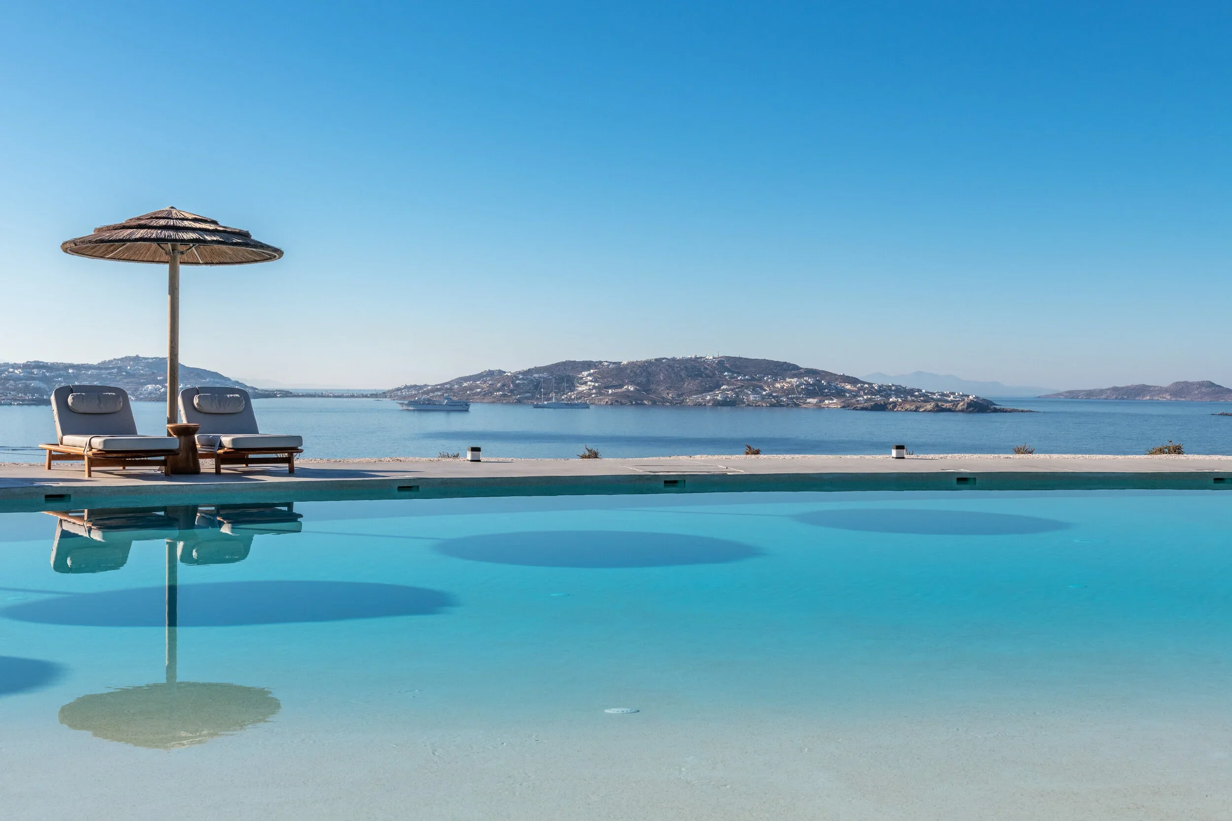 A swimming pool overlooking a large body of water with islands and hills in the background. Two lounge chairs with cushions and an umbrella are positioned on the pool deck.
