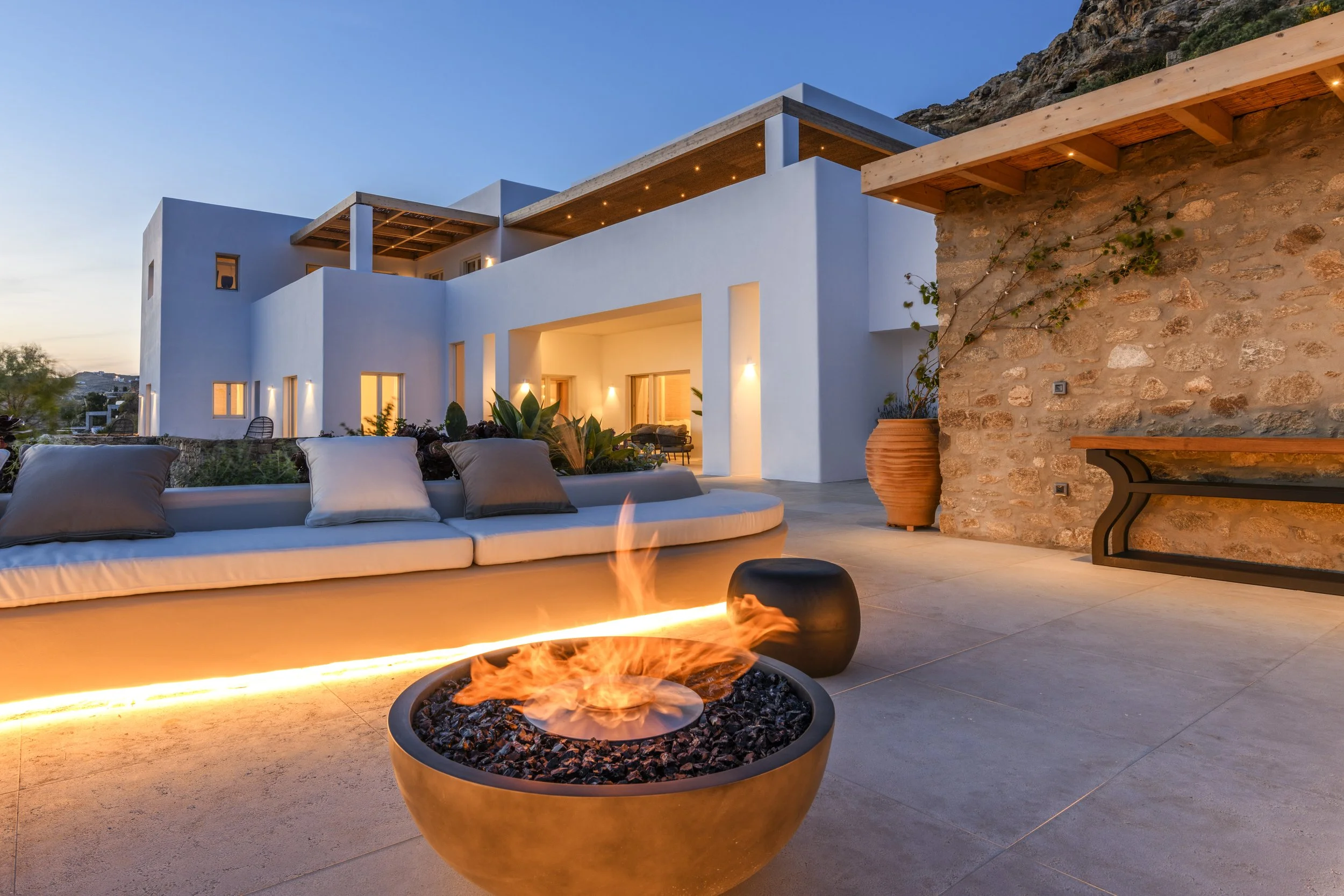 Modern white house with outdoor seating area, fire pit, and stone wall during sunset.