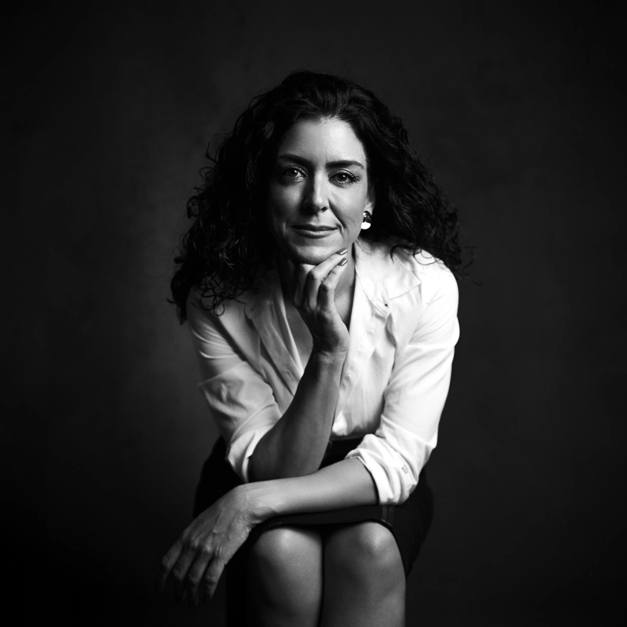 Black and white portrait of a woman with curly hair, wearing a white shirt and dark skirt, sitting with her chin resting on her hand, looking at the camera.