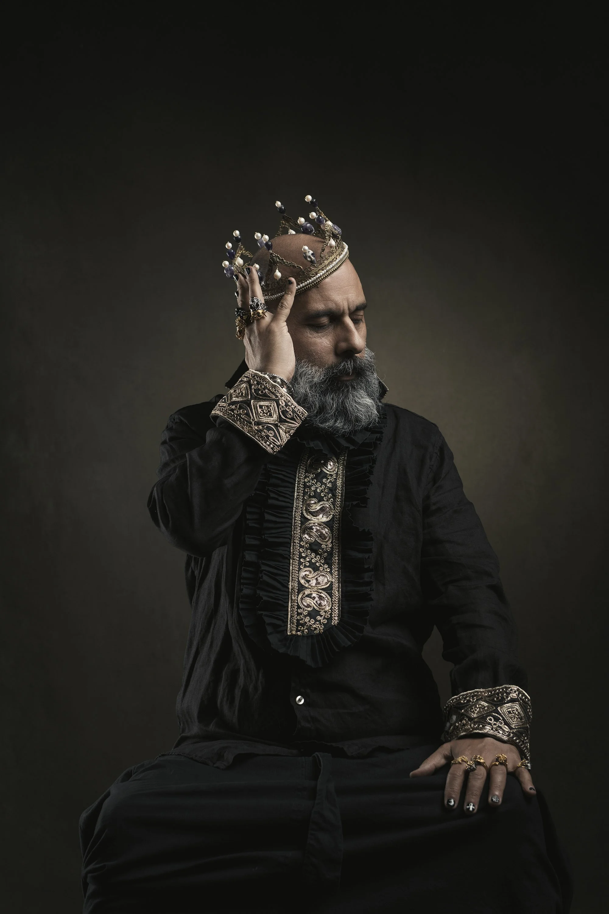 A man with a gray beard, wearing a dark ornate jacket with gold embroidery, is holding a crown with pearls and gemstones near his head in a contemplative pose against a dark background.