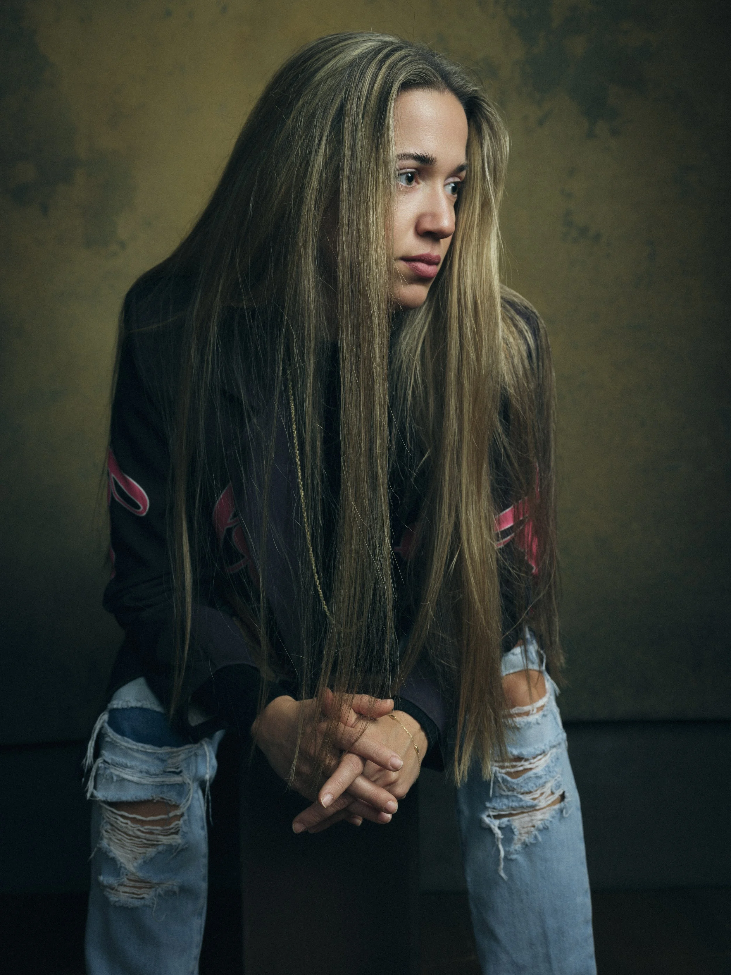 A young woman with long, blonde hair, wearing a dark jacket and ripped jeans, sitting against a textured yellow-green background.