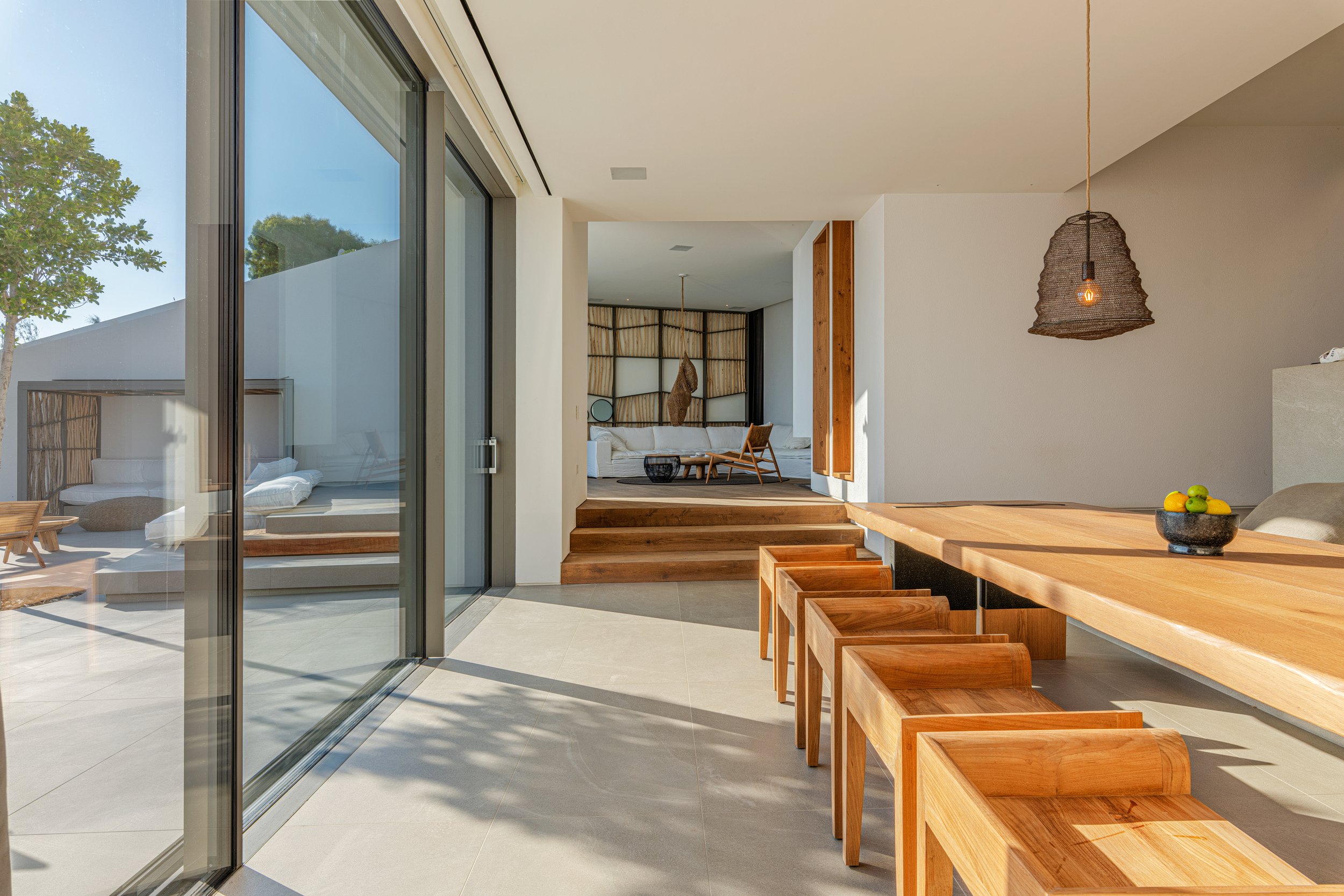 Modern living space with a wooden dining table, chairs, and a sitting area with a sofa and armchair in the background, featuring large sliding glass doors and minimal decor.