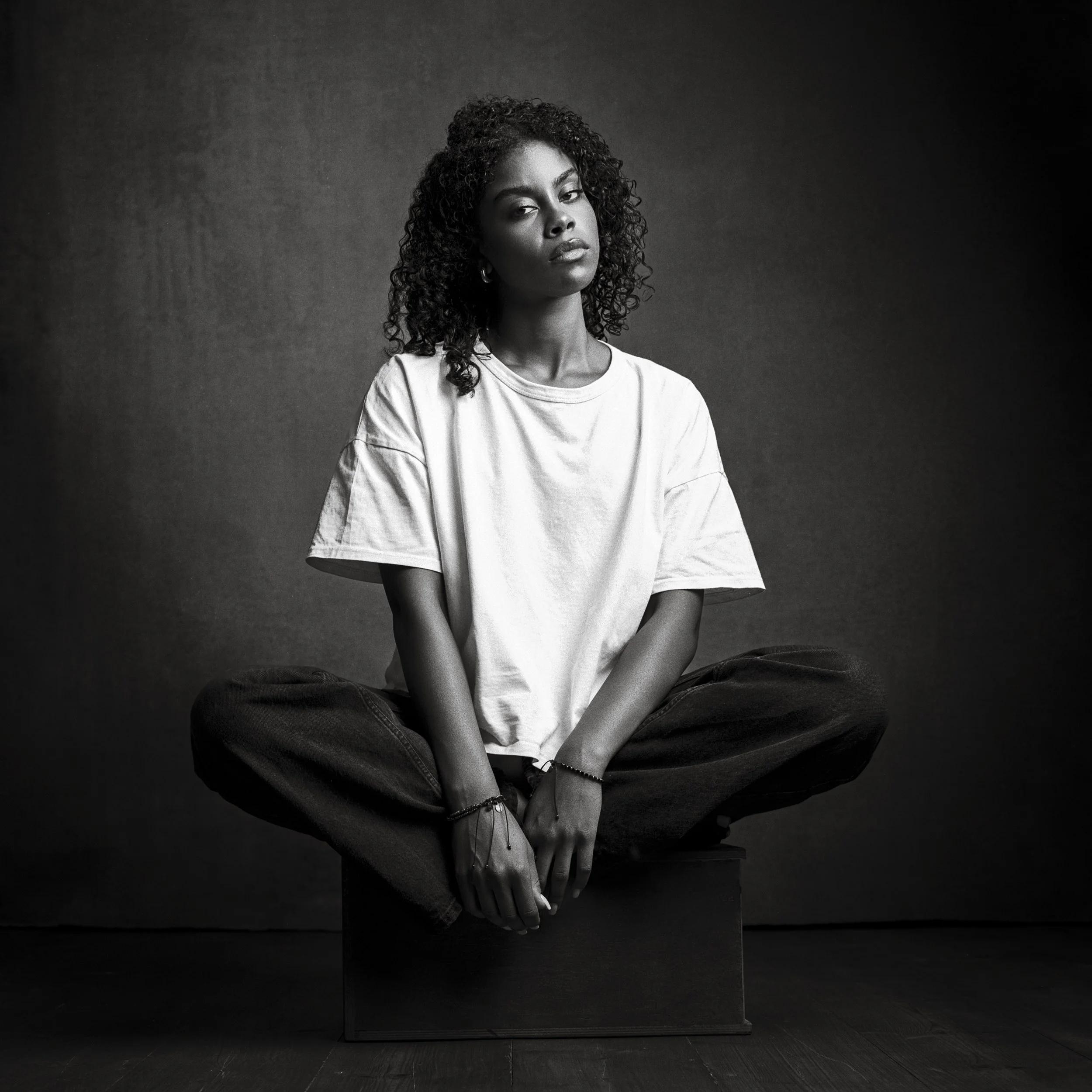 A young woman with curly hair sitting cross-legged on a box, wearing a loose white t-shirt and dark pants, in a monochrome studio portrait.