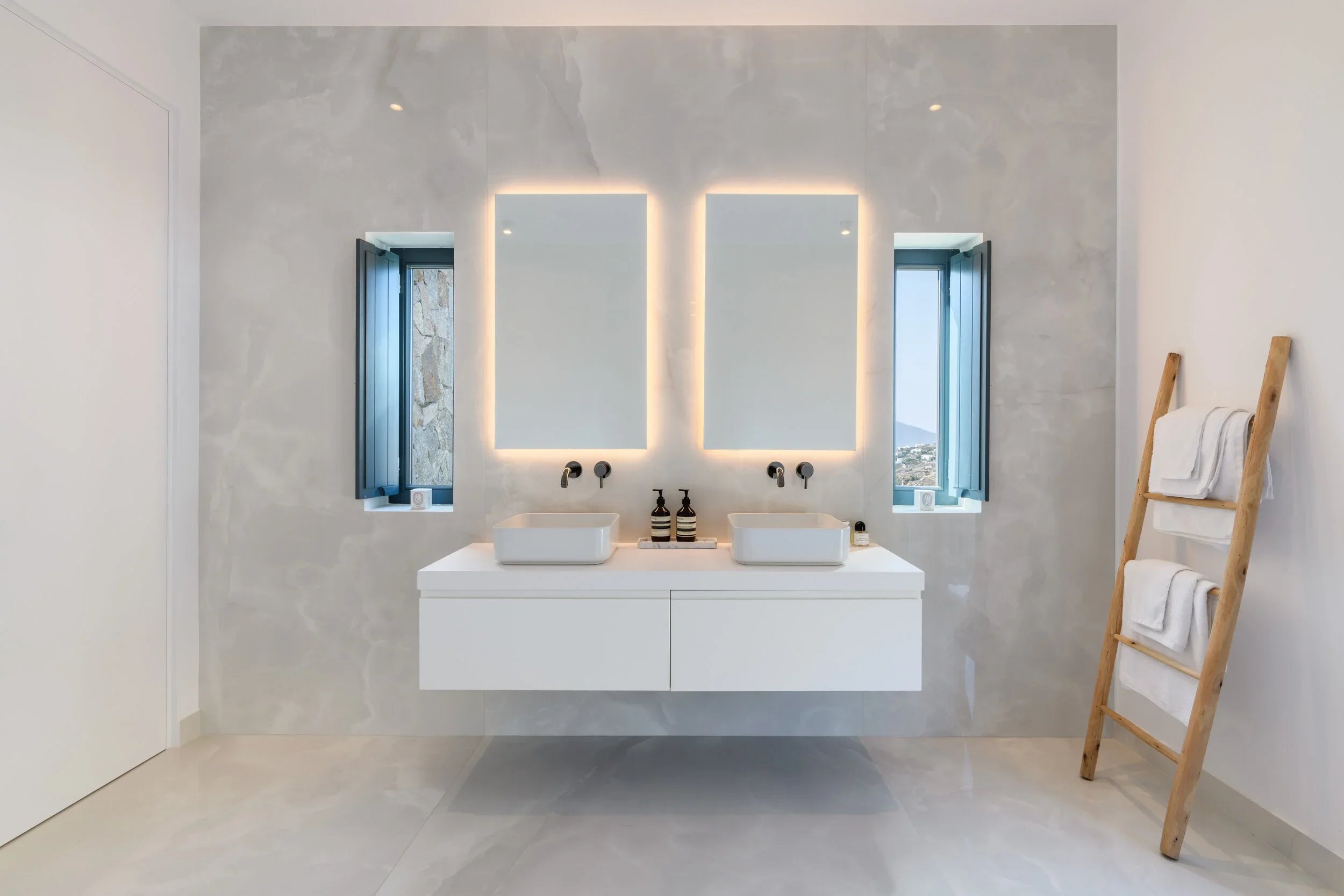Modern bathroom with two rectangular sinks on a floating white vanity, illuminated mirrors, two small windows with blue shutters, and a wooden towel ladder with white towels.