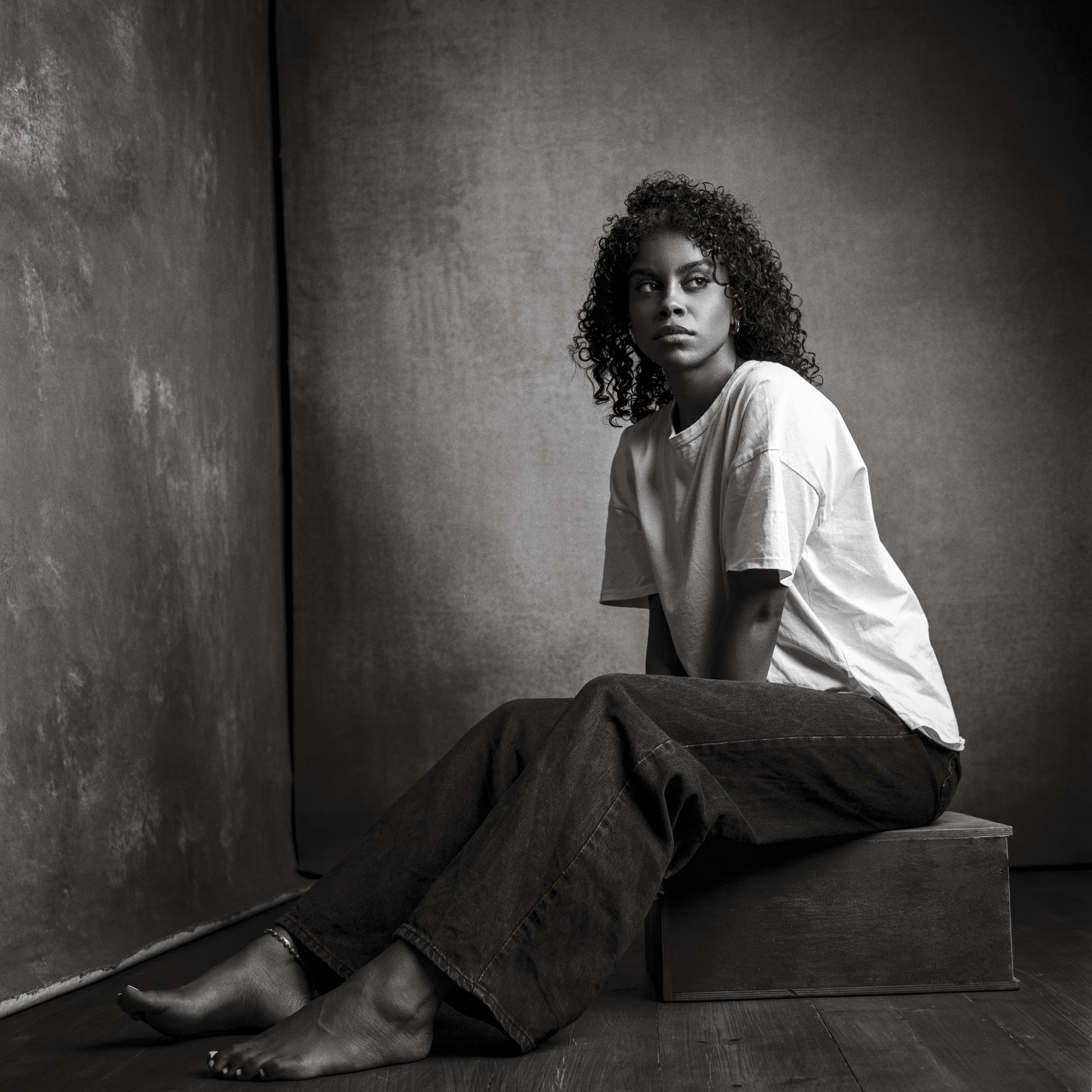 A black and white photo of a woman with curly hair sitting on a wooden block, wearing a loose t-shirt and dark jeans, gazing to the side with a serious expression, against a textured wall background.