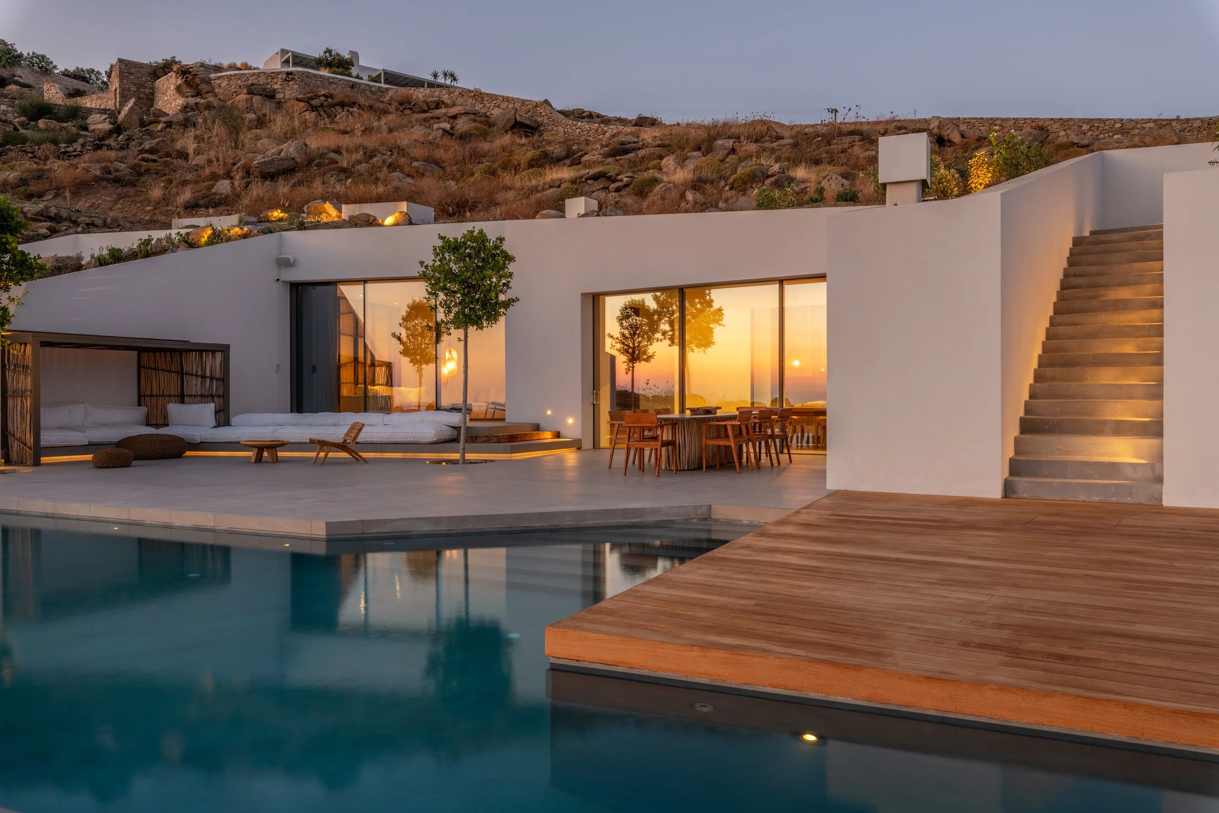 Modern house with large glass sliding doors, outdoor seating, wooden deck, swimming pool, and hillside with rocks and dry bushes at sunset.