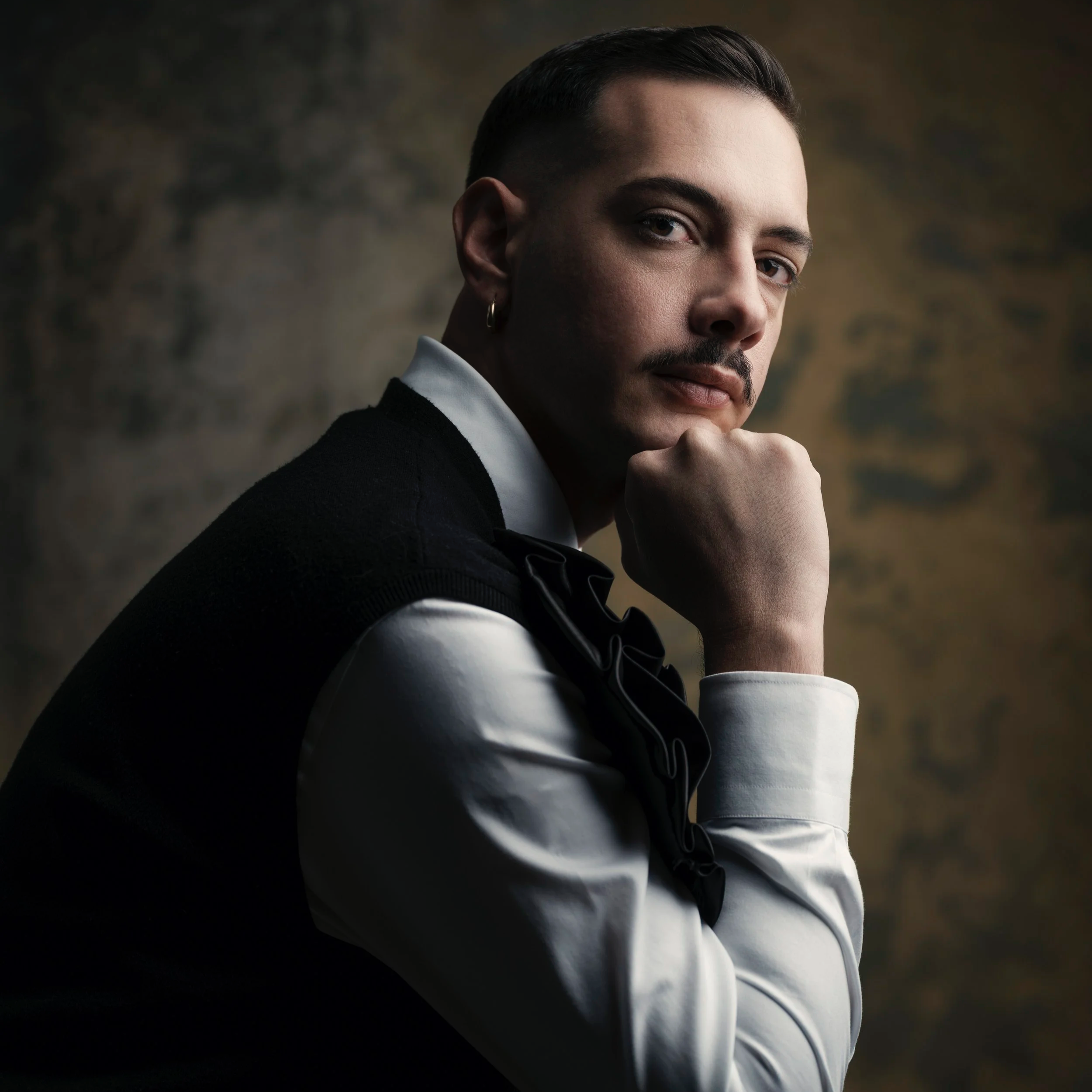 A man with slicked-back hair, mustache, and a small hoop earring in his left ear, resting his chin on his hand, dressed in a black vest over a white shirt with a bow tie, looking thoughtfully at the camera against a dark, textured background.