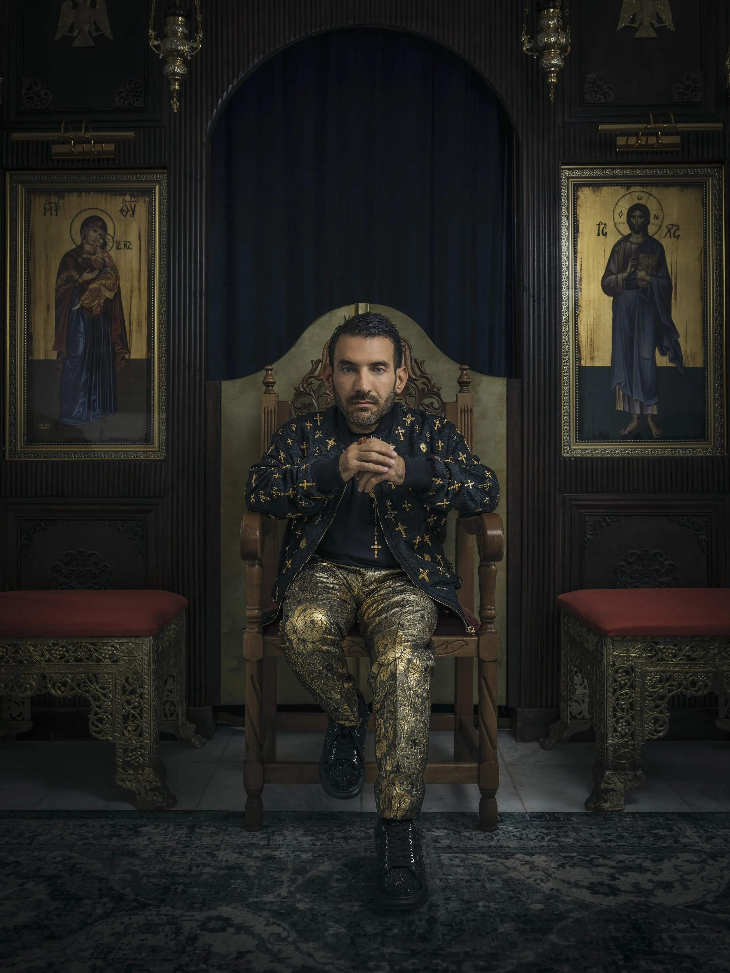 A man sitting on a throne-like chair in a room with religious icons on the walls, wearing a dark jacket with gold crosses and patterned gold pants.