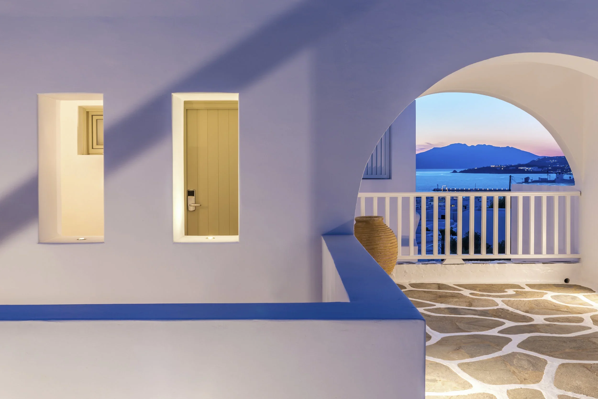 A terrace with white walls and a stone floor, featuring three illuminated windows and a door, and a view of a coastal landscape with mountains, water, and a sunset in the background.