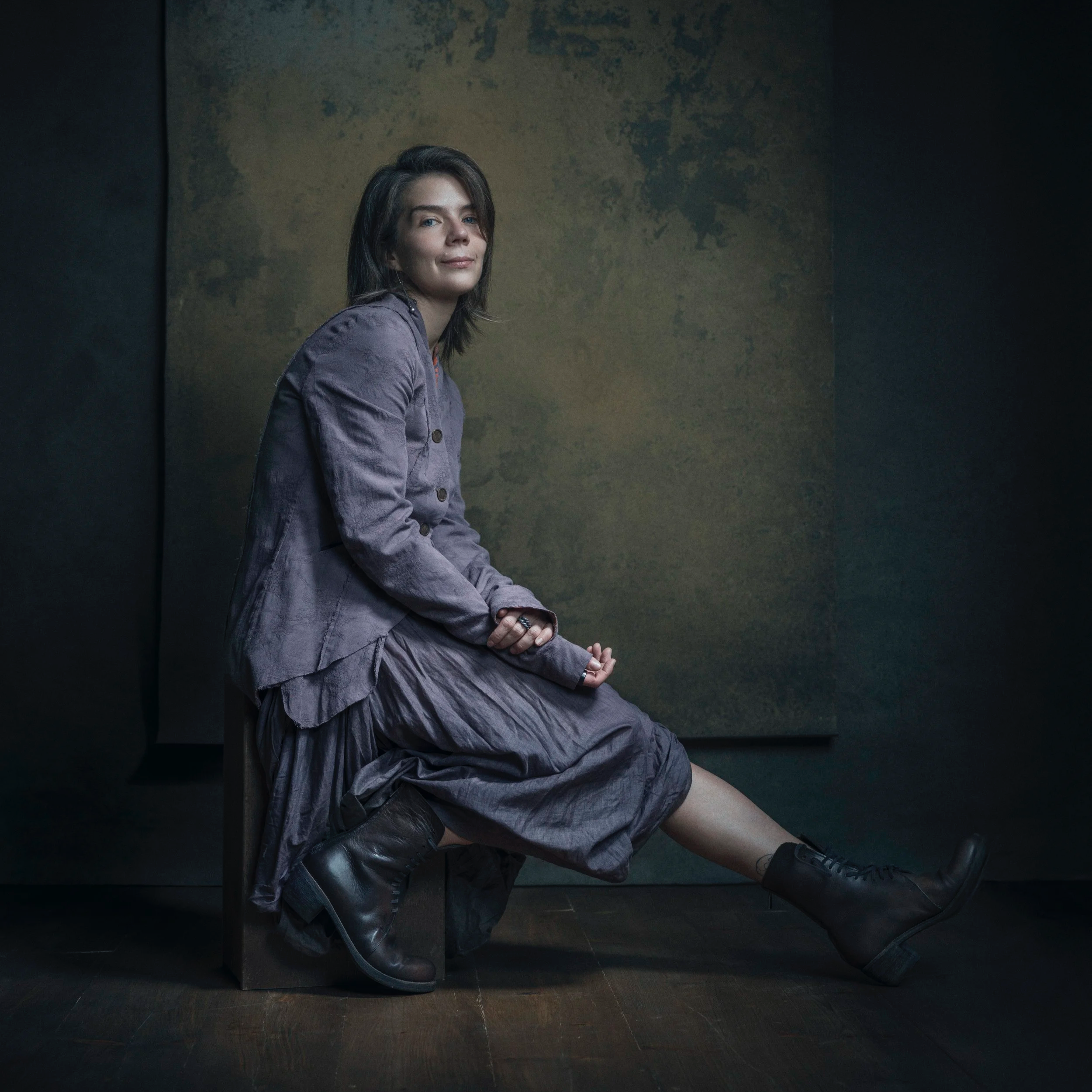 Woman sitting on a wooden block against a dark and textured background, wearing a purple dress, a purple jacket, and black combat boots, with short dark hair, looking at the camera with a slight smile.