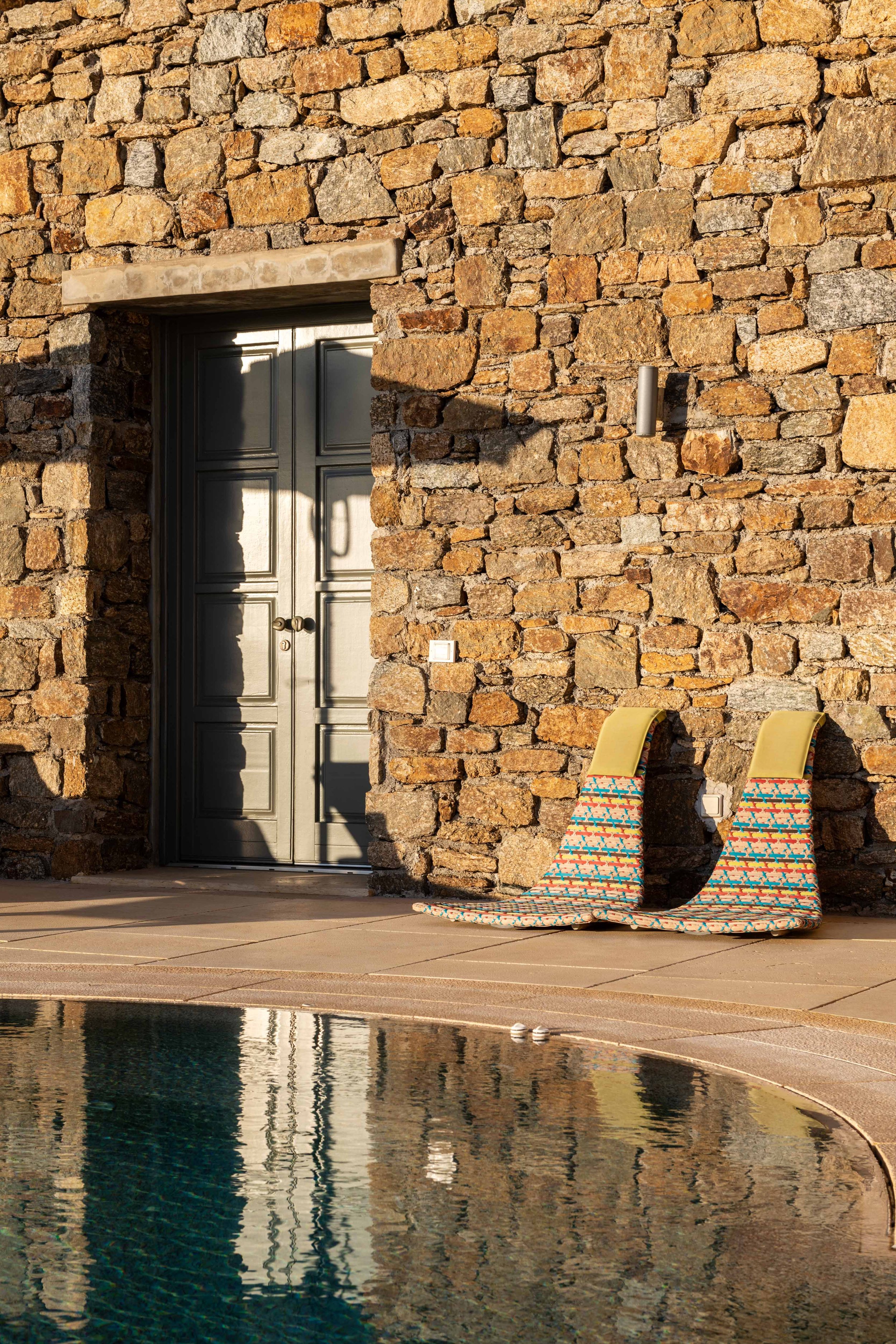 A stone wall with a closed gray door, two lounge chairs with colorful striped cushions, and a swimming pool reflecting the wall and door.