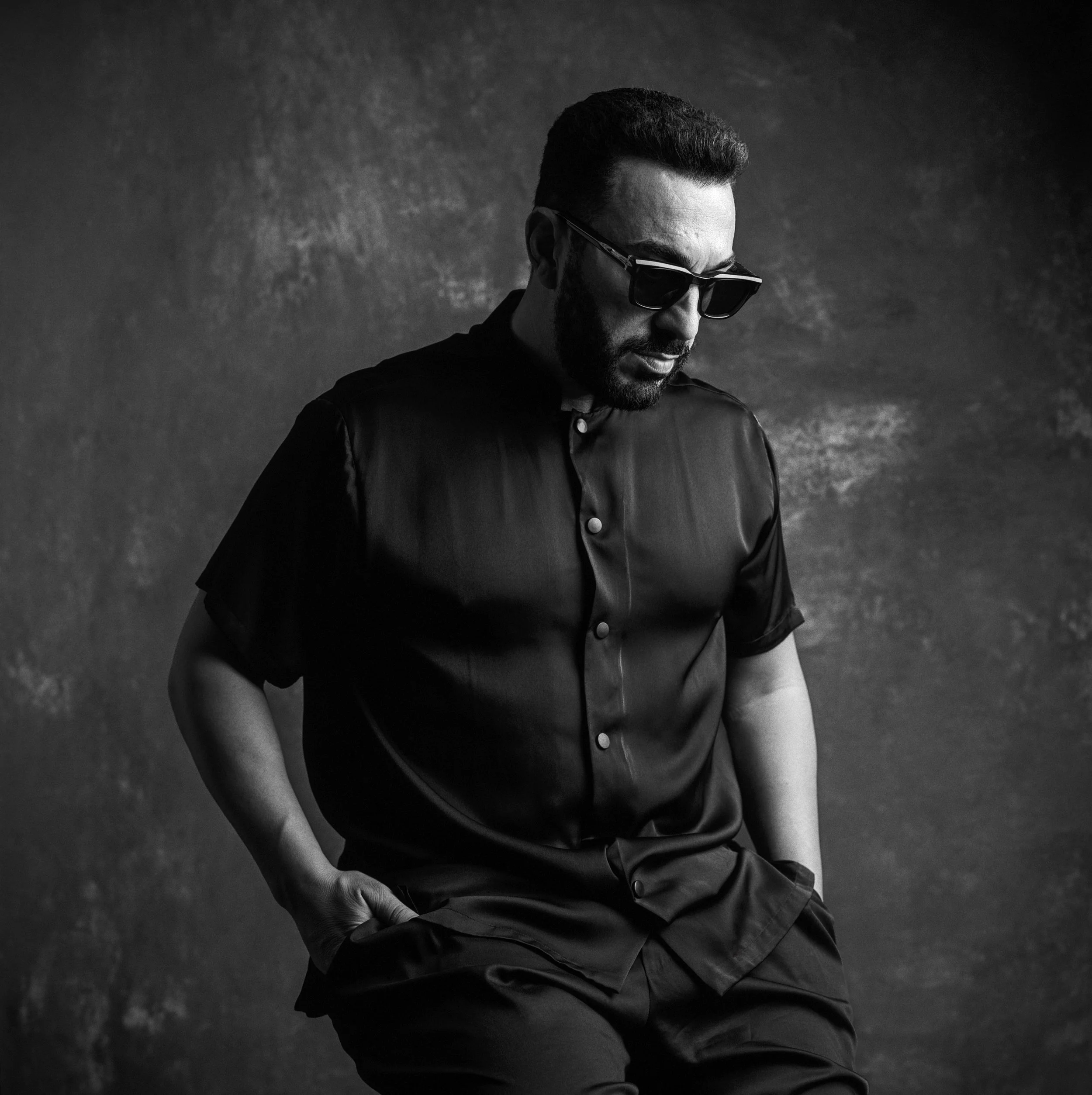Black and white portrait of a man wearing sunglasses, a short-sleeved button-up shirt, and pants, standing against a textured backdrop. He has a short hairstyle and beard, with his hands in his pockets, looking down.