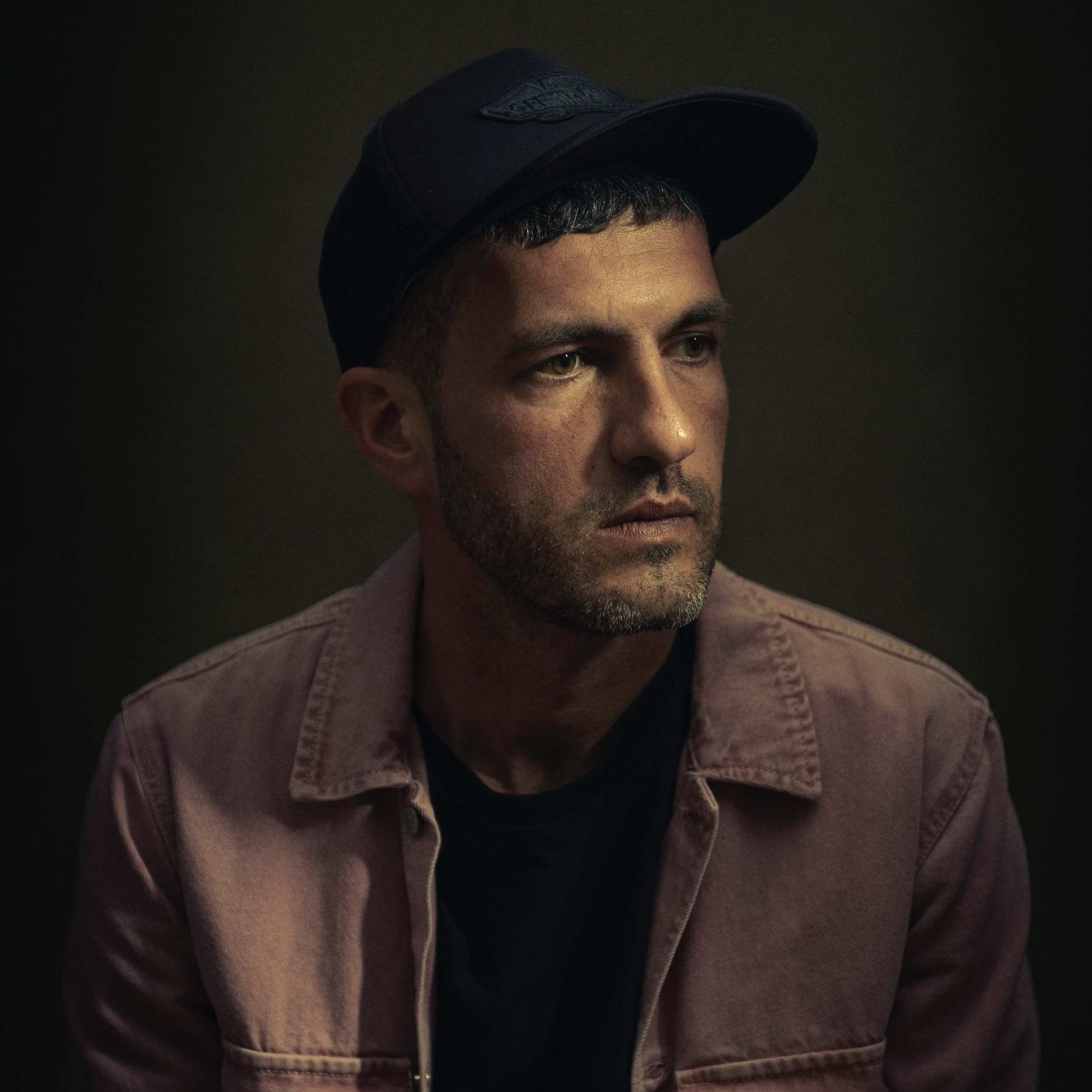 portrait of a man with a short beard wearing a black cap and a brown jacket, looking to the side against a dark background.