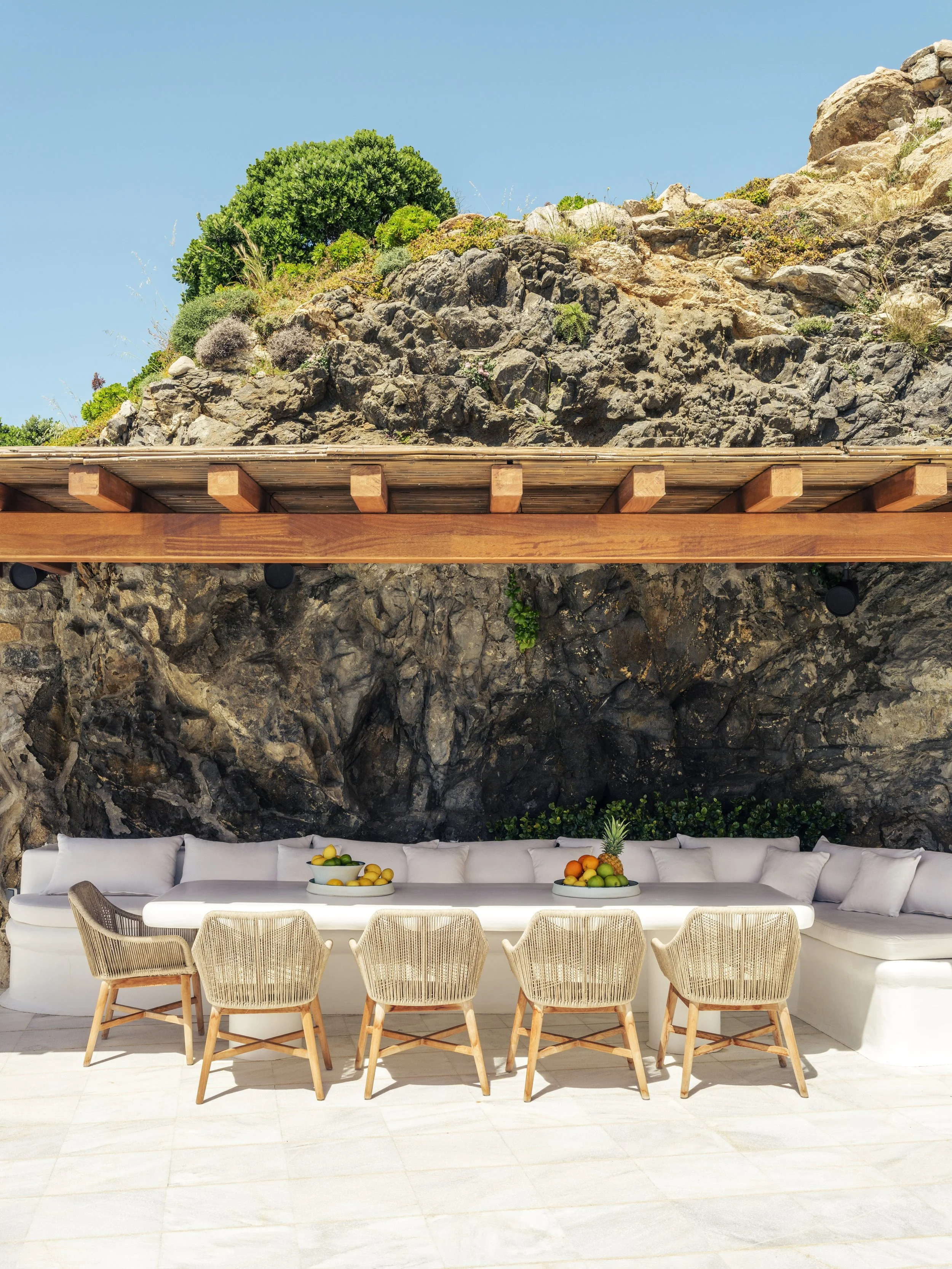 Outdoor patio with a curved white sofa, six wicker chairs, a white table with bowls of fruit, set against rock walls and a rocky hillside with green vegetation under a blue sky.