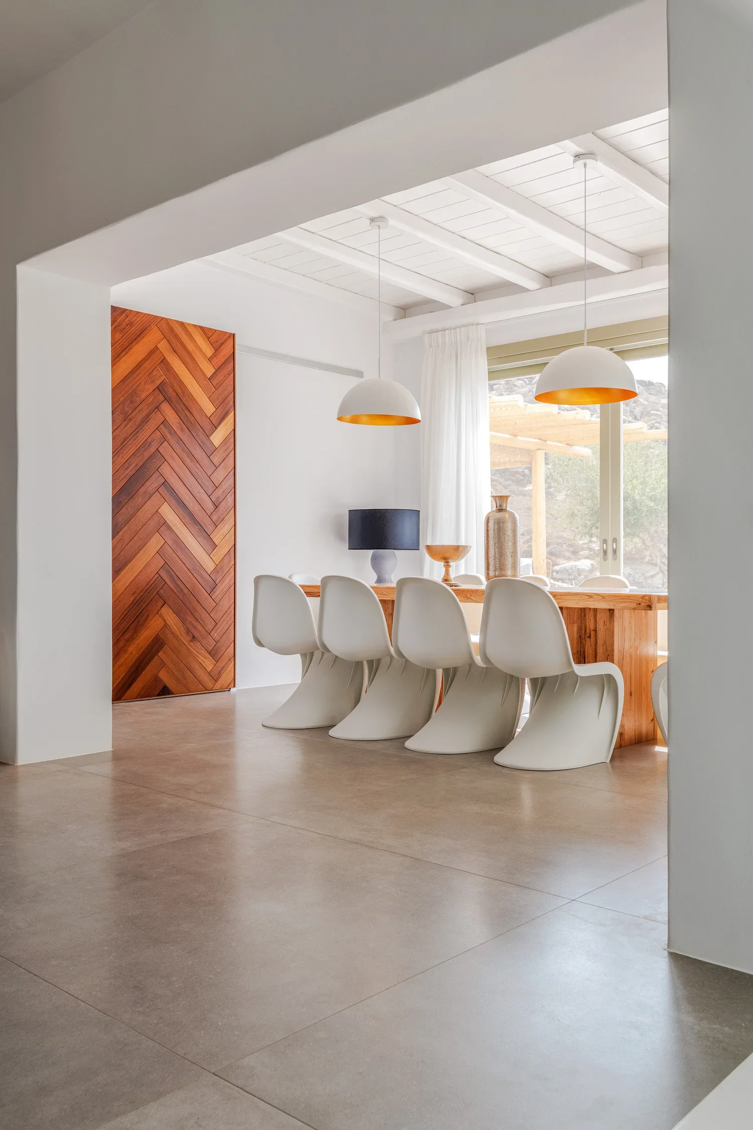 A modern dining room with a wooden table, six white chairs, two pendant lights hanging above, a window with white curtains, and decorative vases on the table.