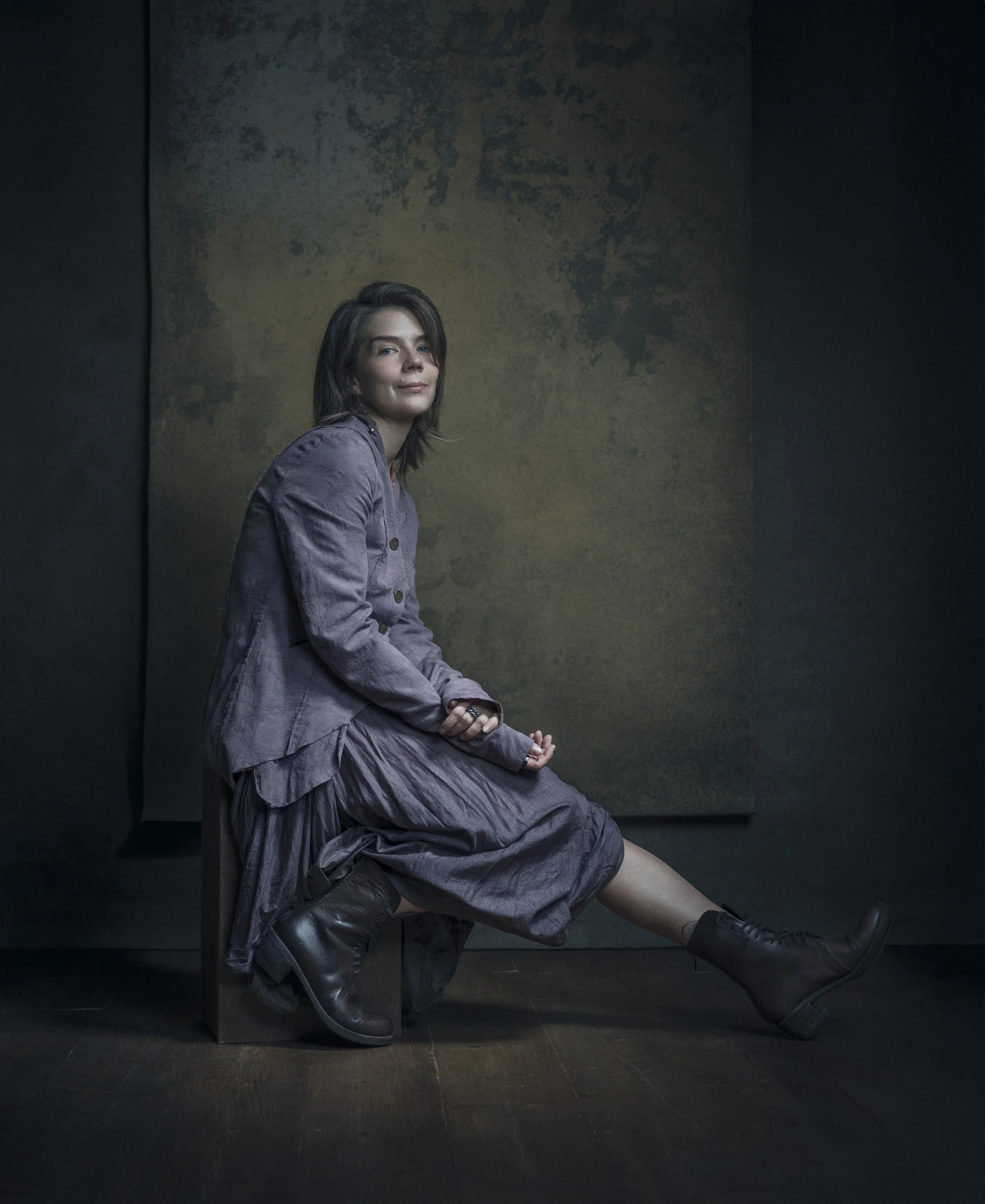 A woman sits sideways on a black chair against a dark wall with a textured framed artwork behind her. She has shoulder-length brown hair and is wearing a purple jacket, a gray pleated skirt, and black boots, with her hands resting on her lap.