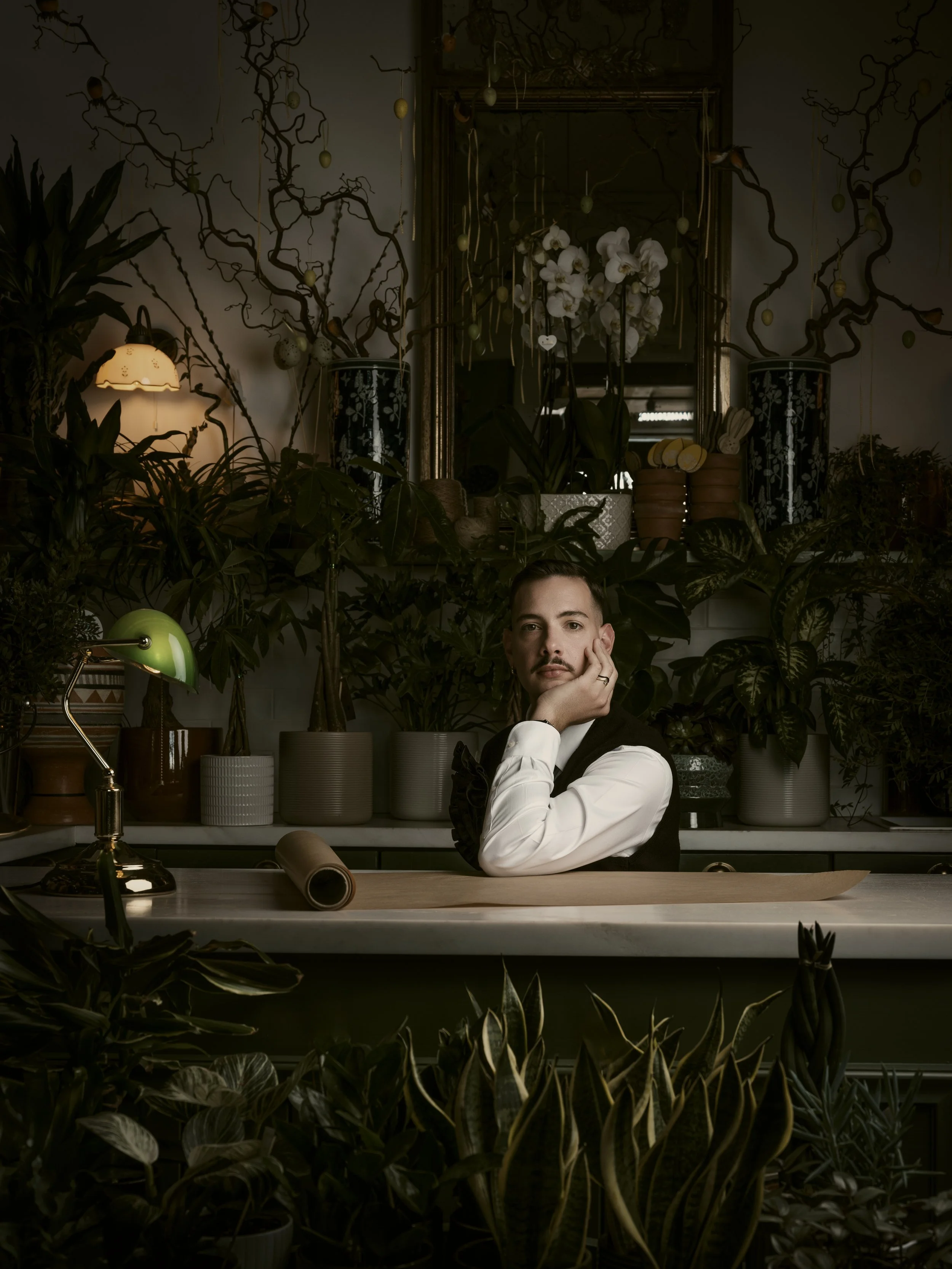 A man sitting at a desk in a dimly lit room with plants and decorative items, resting his chin on his hand and looking at the camera.