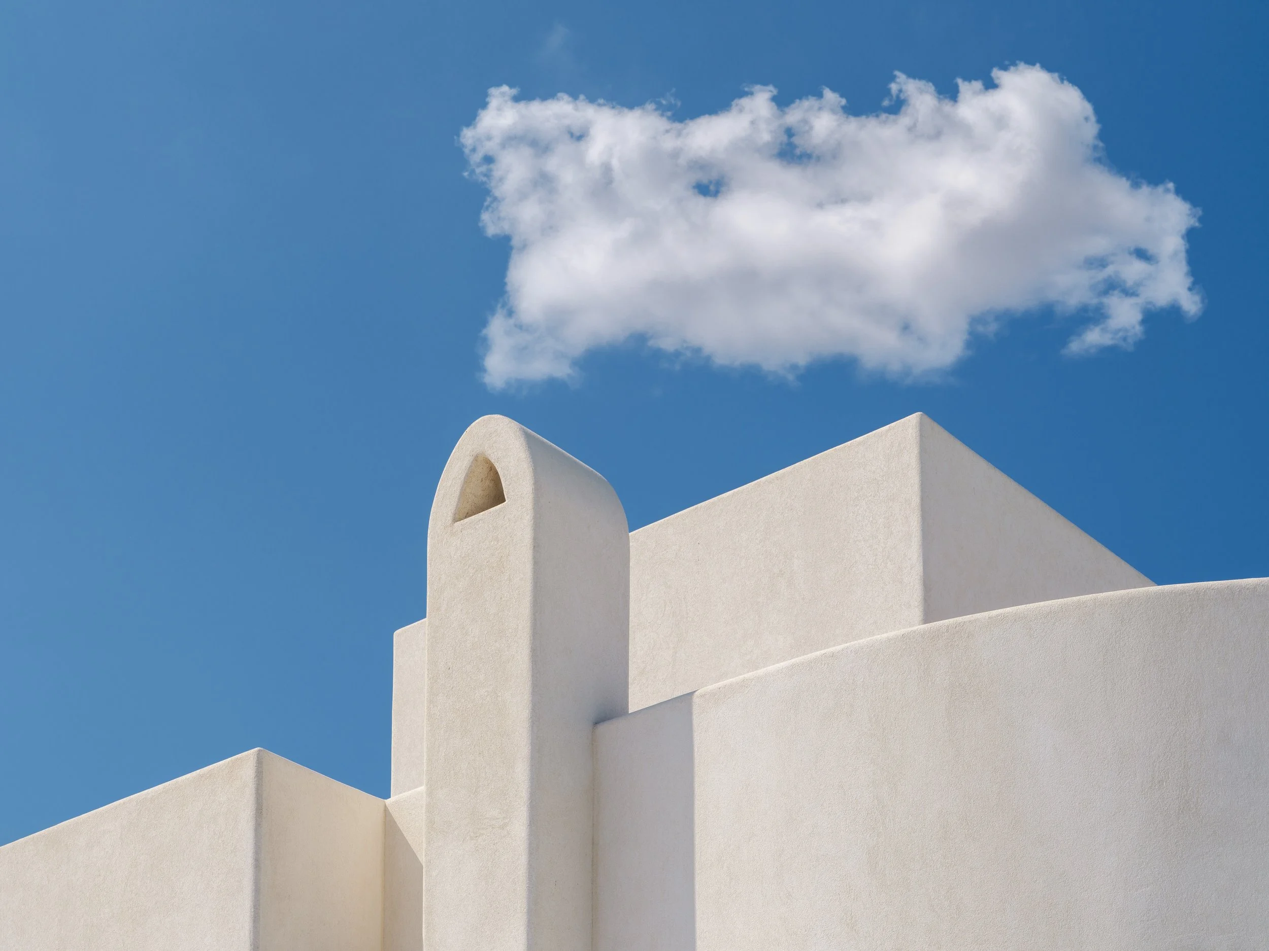 A white building with smooth curved and straight architectural features under a clear blue sky with a single fluffy white cloud.