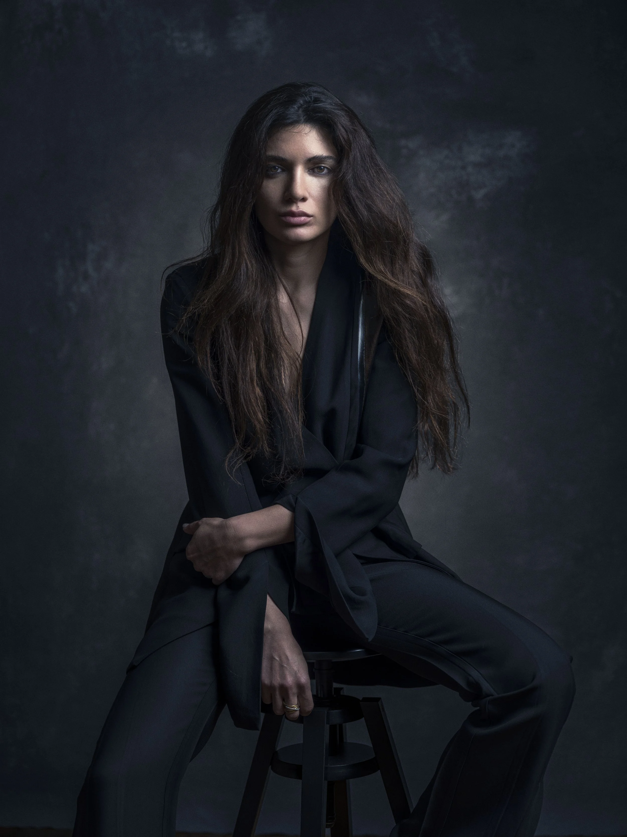 A woman with long, wavy brown hair wearing a black blazer and black pants sitting on a stool against a dark background.