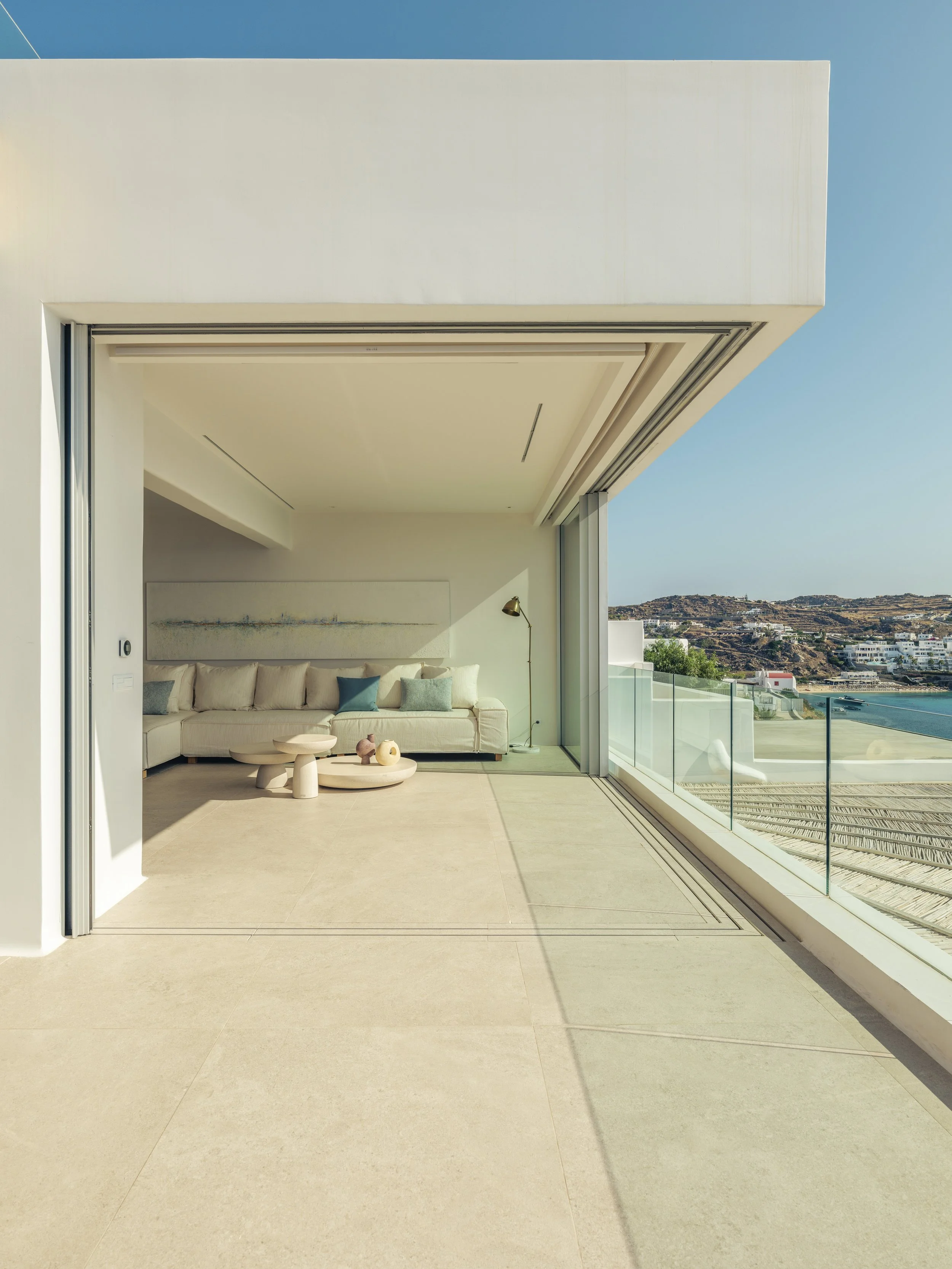 Modern open-concept living room with a large white sectional sofa, abstract wall art, and a floor lamp, opening to a balcony with ocean and landscape views.