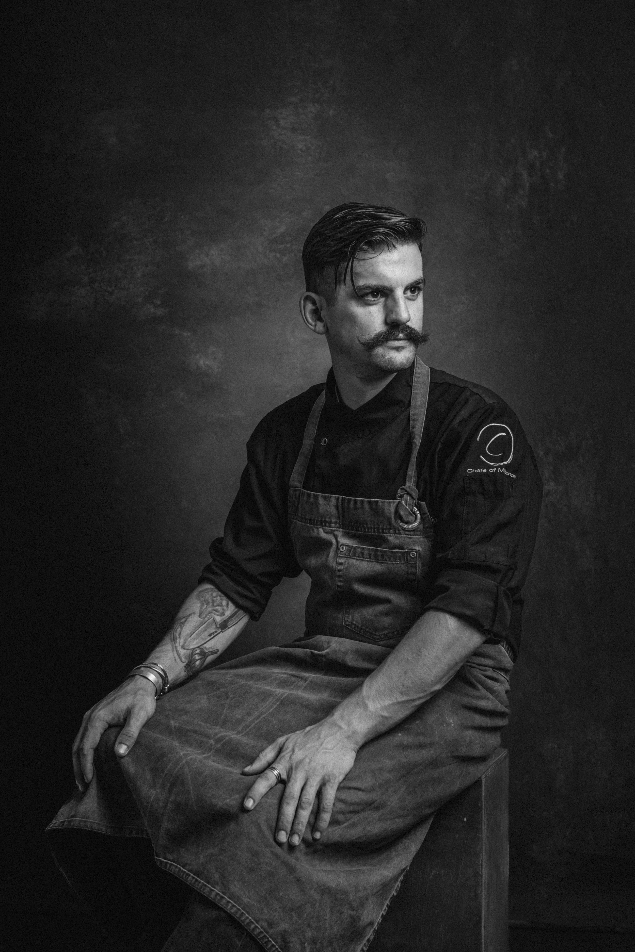 Black and white portrait of a man with a mustache and styled hair, wearing a chef's jacket and apron, sitting on a stool against a dark background.
