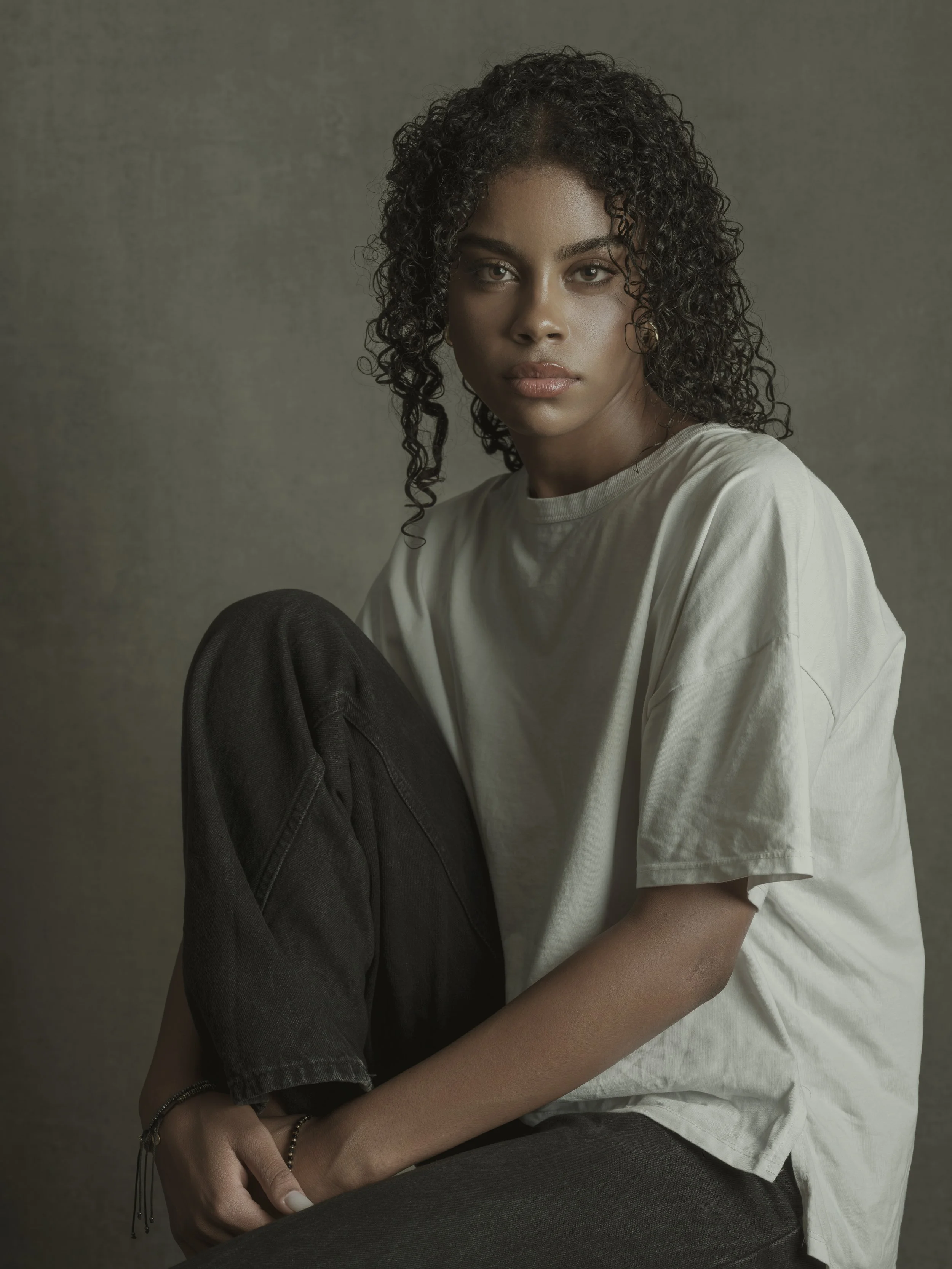 A young woman with dark curly hair, wearing a loose white t-shirt and black pants, sitting with one knee up and looking at the camera against a plain background.