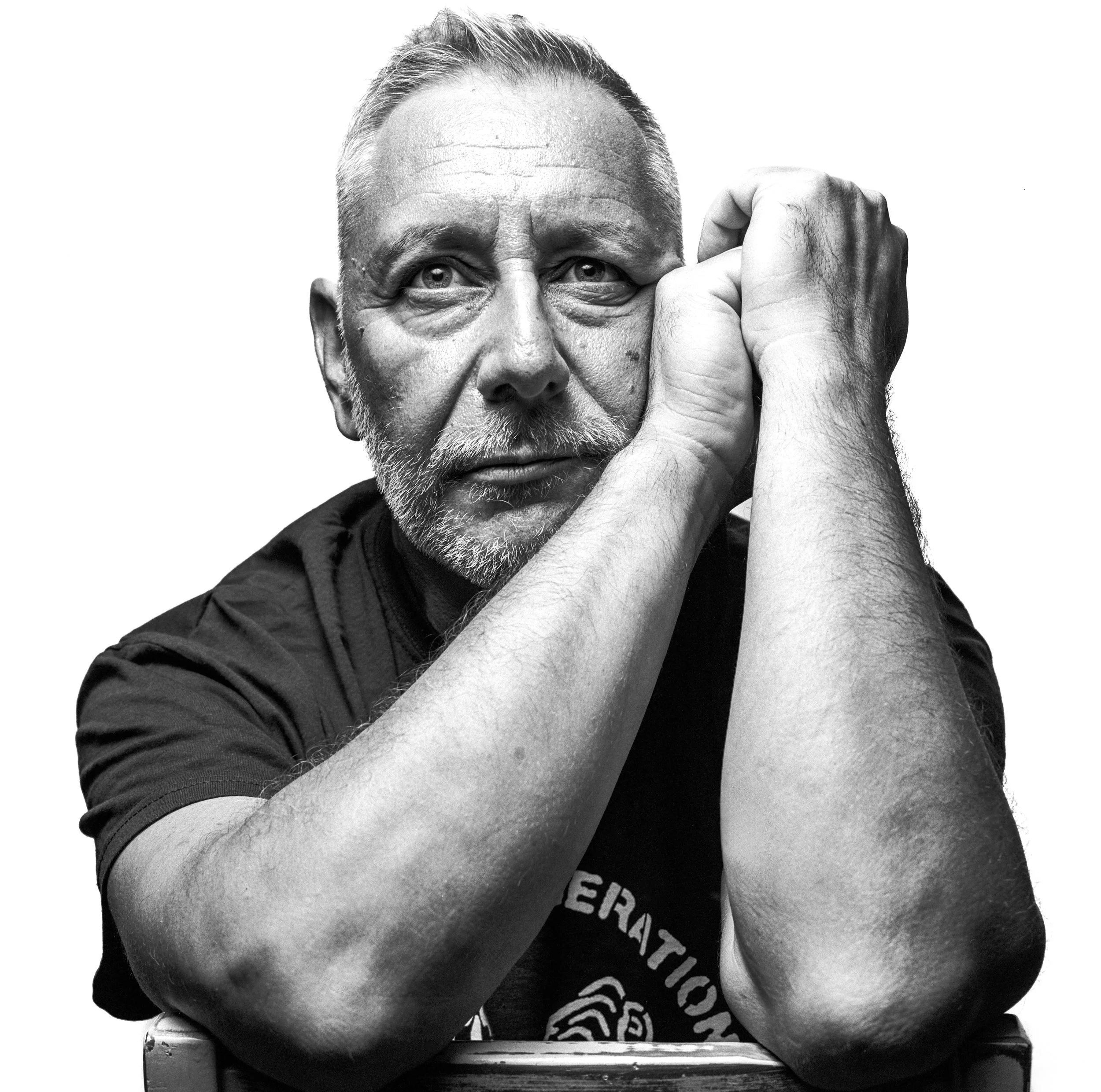 A black and white close-up of an older man with a beard and short hair, resting his head on his folded arms, looking thoughtfully.