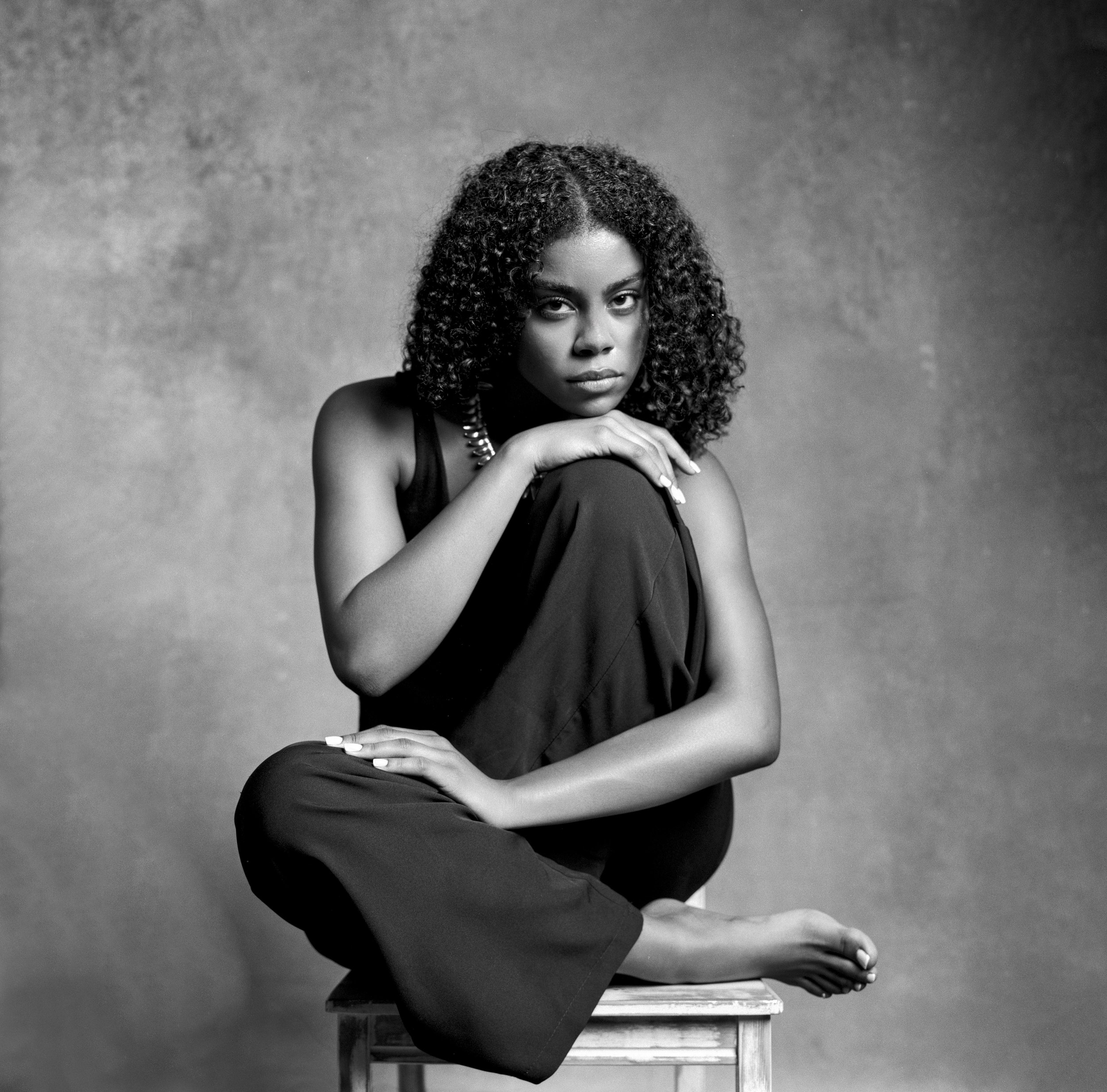 A black and white portrait of a young woman with curly hair, sitting on a small stool, wearing a sleeveless top and pants, with one arm resting on her knee and the other hand touching her chin, looking directly at the camera.
