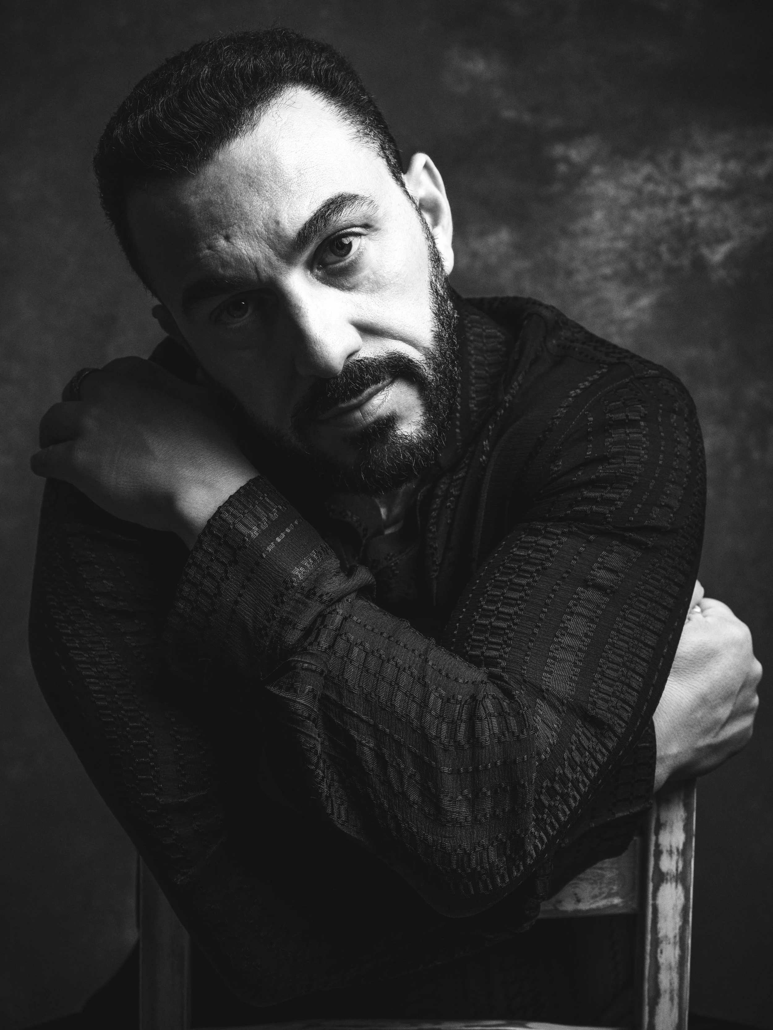 Black and white portrait of a man with a beard and mustache, looking at the camera with a thoughtful expression, sitting on a chair with one arm resting on the back.