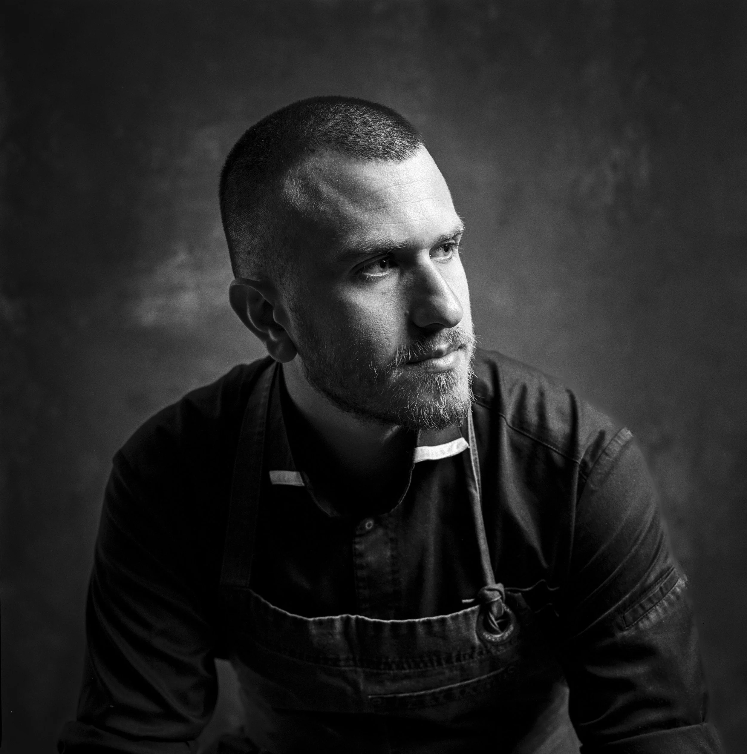 Black and white portrait of a man with a beard and short hair, looking to the side, wearing a dark shirt and apron against a dark textured background.