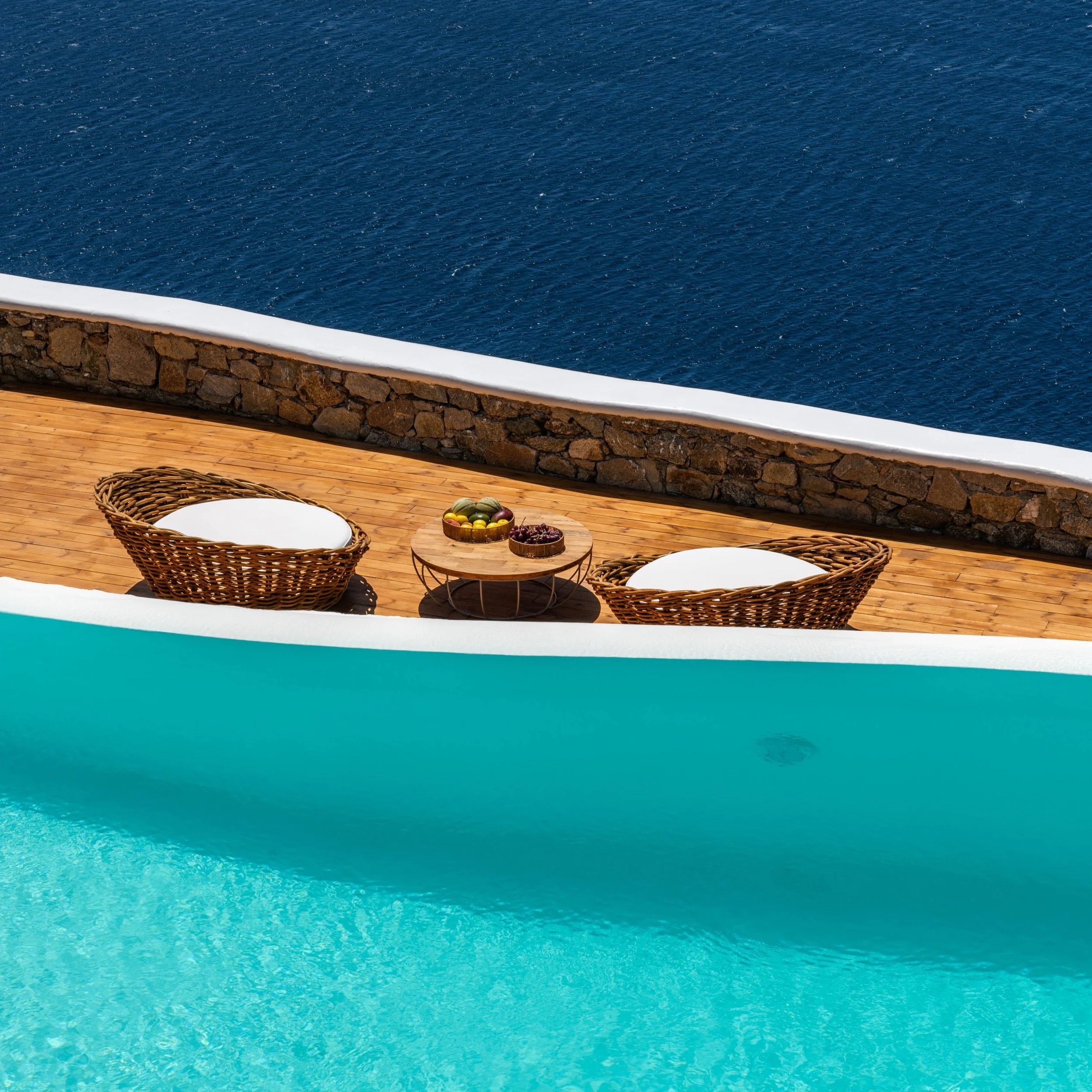 Poolside view with two wicker lounge chairs and a small round table with bowls of fruit, overlooking the ocean on a sunny day.