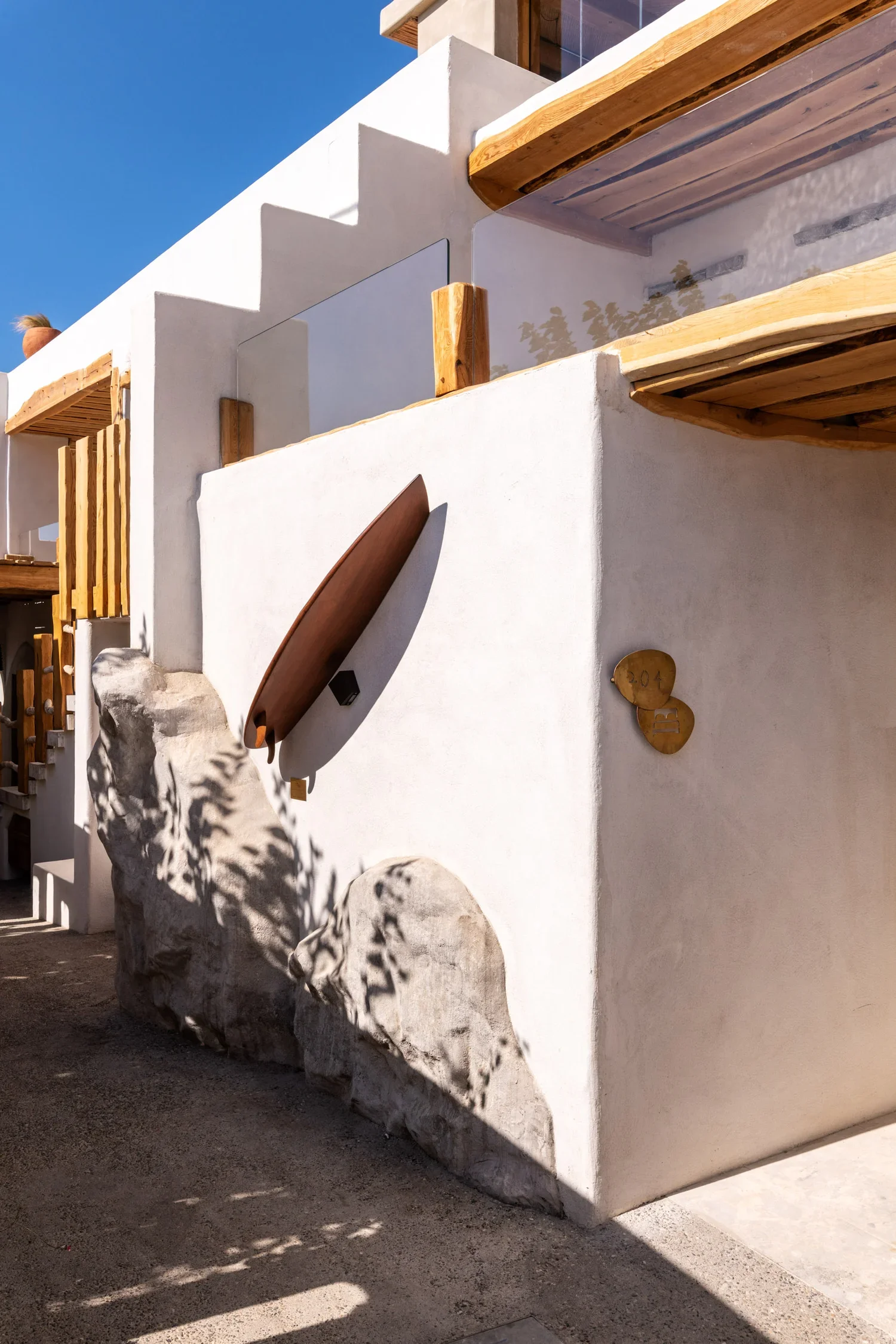 A modern white building with wooden balcony accents, a surfboard mounted on the wall, and shadows of trees cast on the wall. The sky is clear and blue.