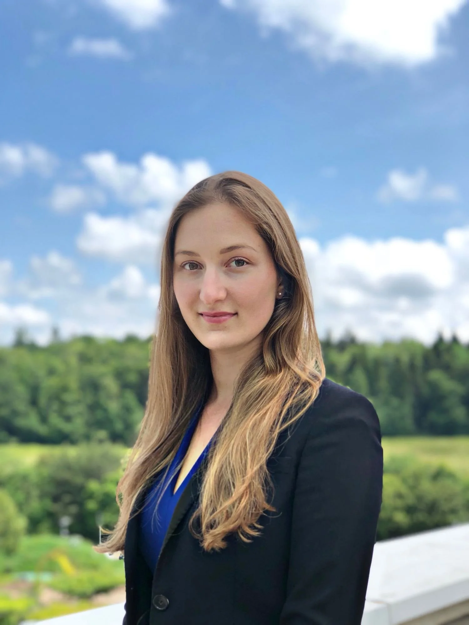 Portrait photo of Maria Matskevich, a freelance B2B content marketer. Image show a woman with long, light brown hair wearing a dark blazer and blue blouse, standing outside against a backdrop of green trees, blue sky, and white clouds.