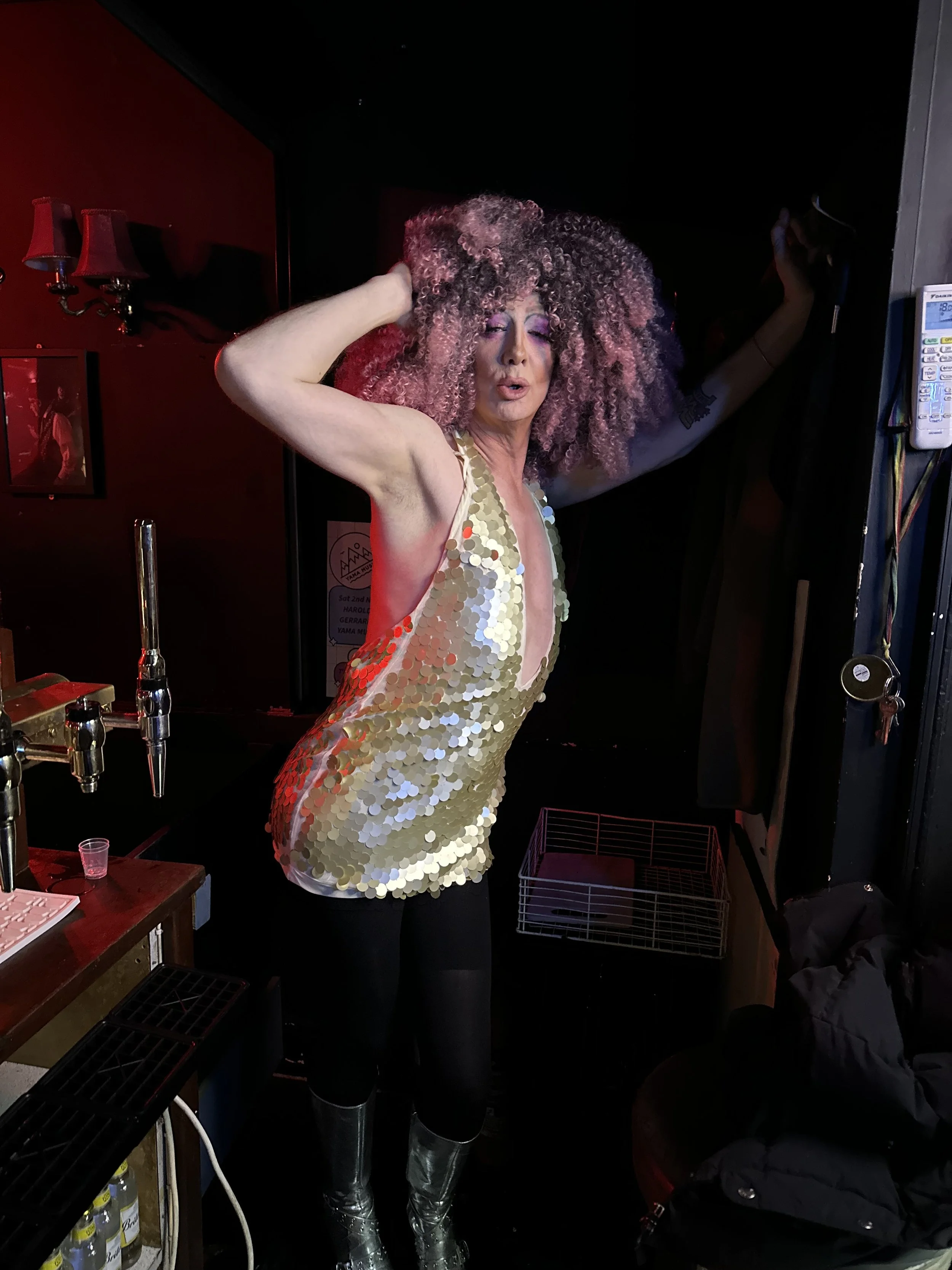 A drag queen with voluminous curly hair wearing a shiny gold sequin dress, posing with one hand in her hair and the other raised, in a dimly lit bar or club.