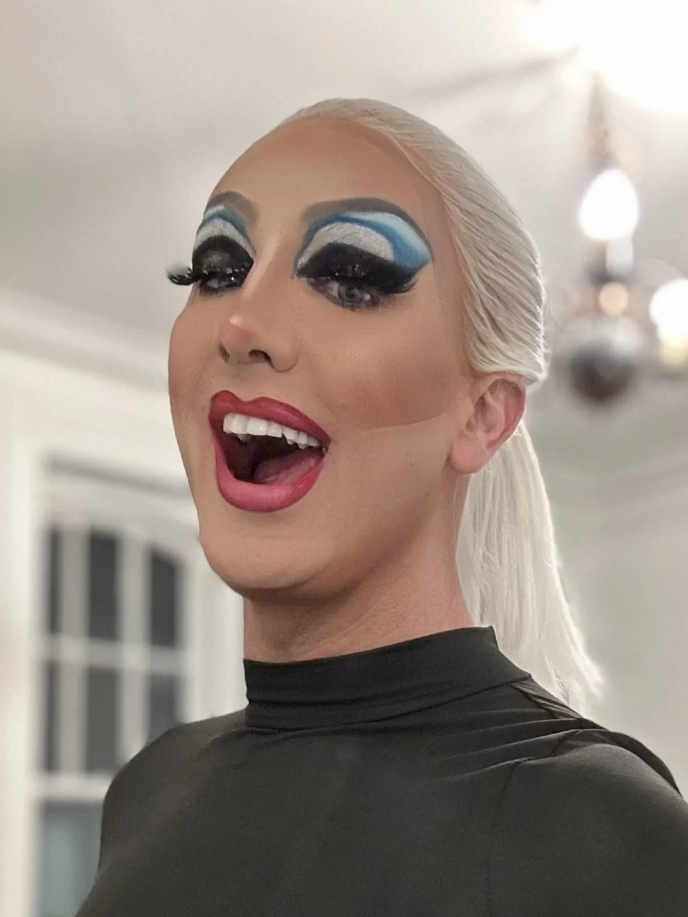 A person with platinum blonde hair styled back, wearing dramatic eye makeup with blue and silver eyeshadow, thick false eyelashes, bright red lipstick, and a black top, smiling and showing teeth with an open mouth.