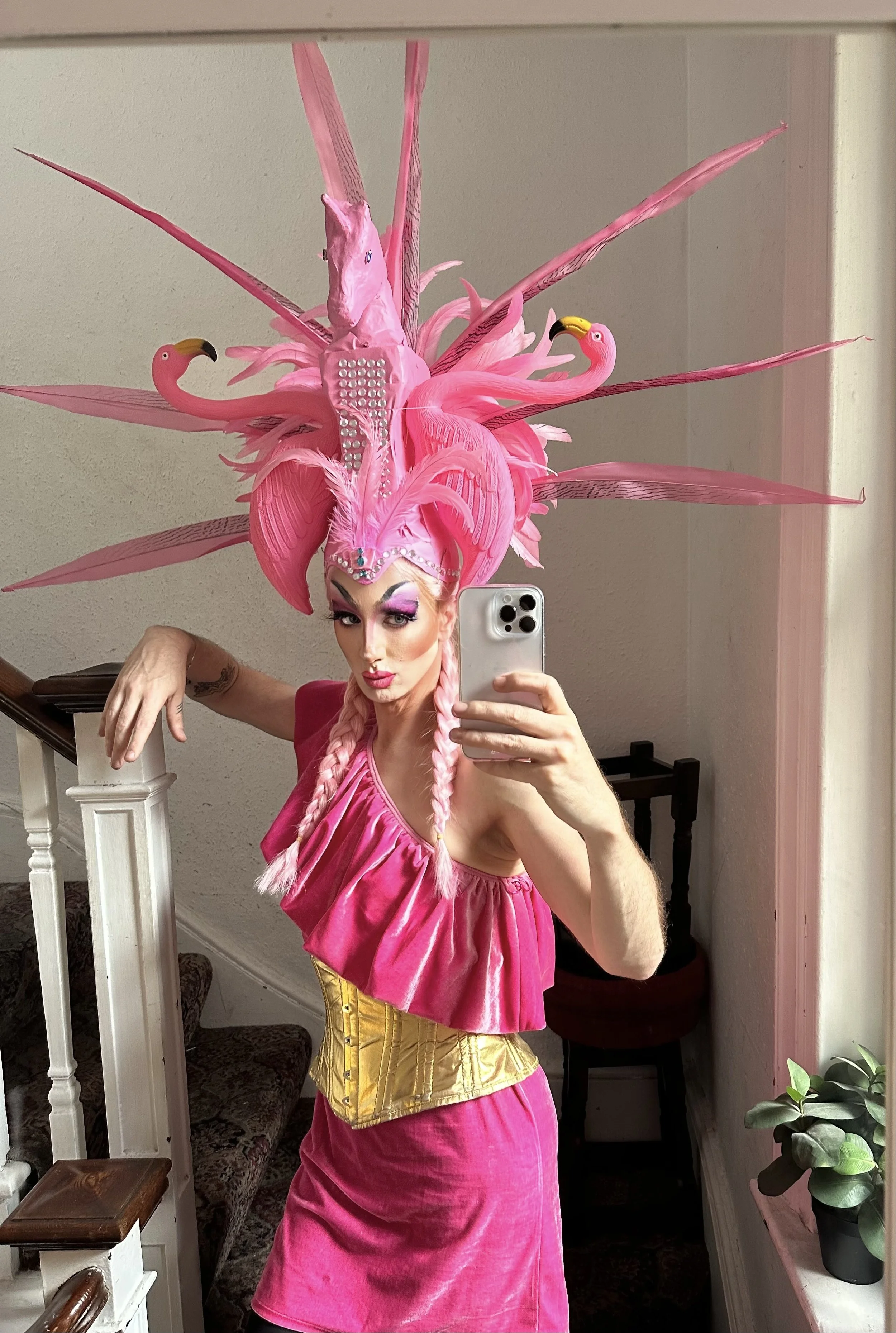 Person taking mirror selfie dressed in vibrant pink outfit with elaborate pink feathered headdress adorned with flamingo and unicorn shapes, and a yellow corset.