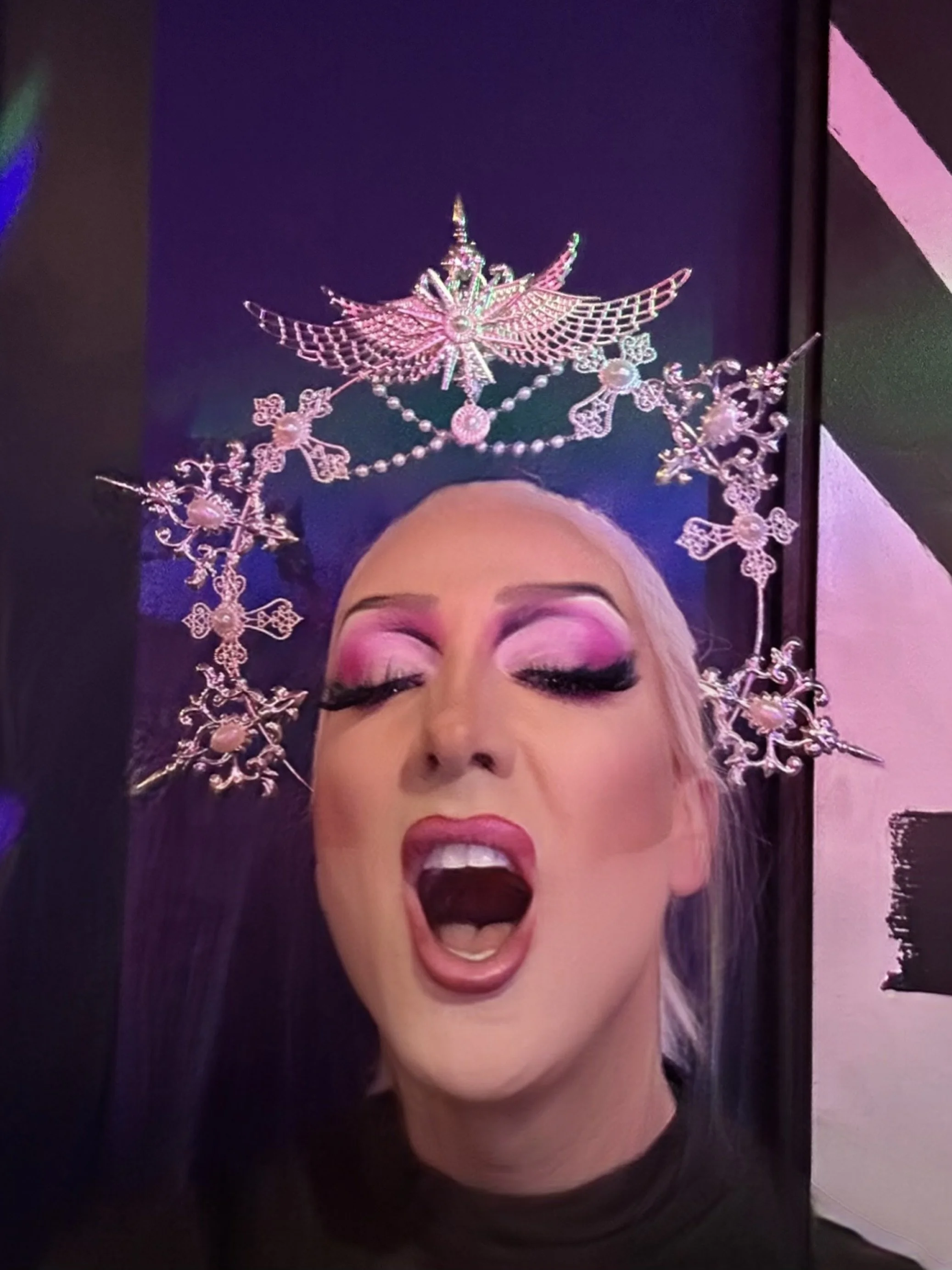 A person with makeup and long false eyelashes wearing a headpiece made of pink and silver snowflakes and angels, with their eyes closed and mouth open as if singing or shouting.