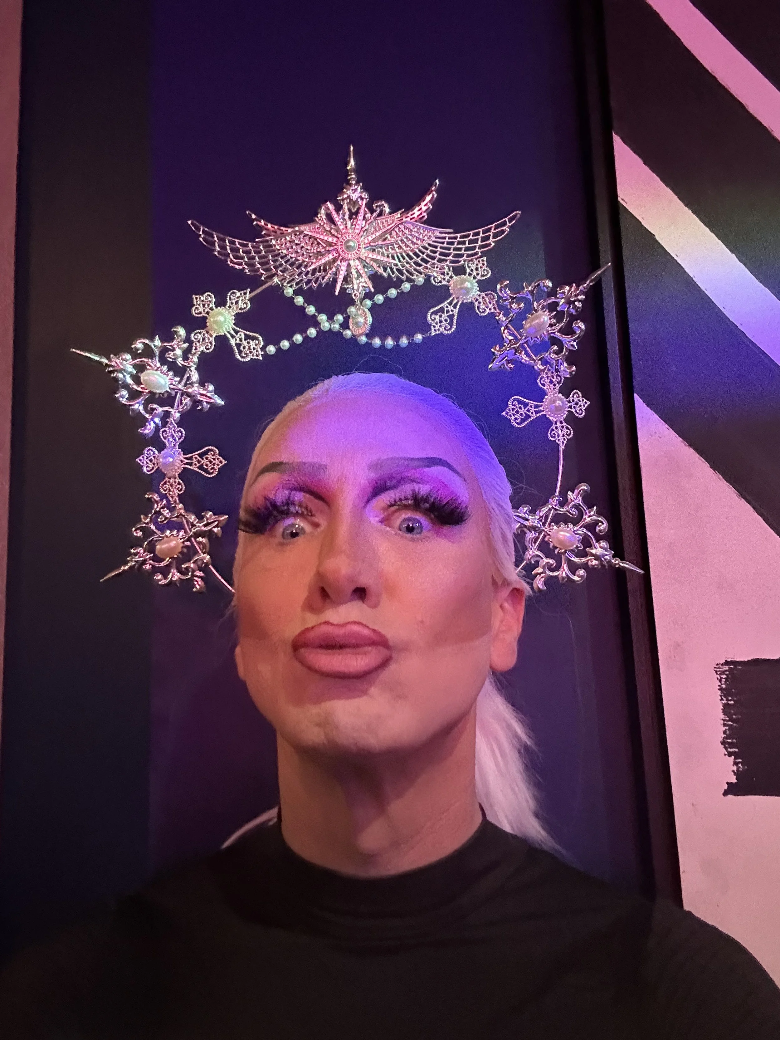 Person with platinum blonde hair wearing outrageous makeup and a headpiece resembling a metallic star, pearls, and wings, taking a selfie in front of a dark background with colorful lighting.